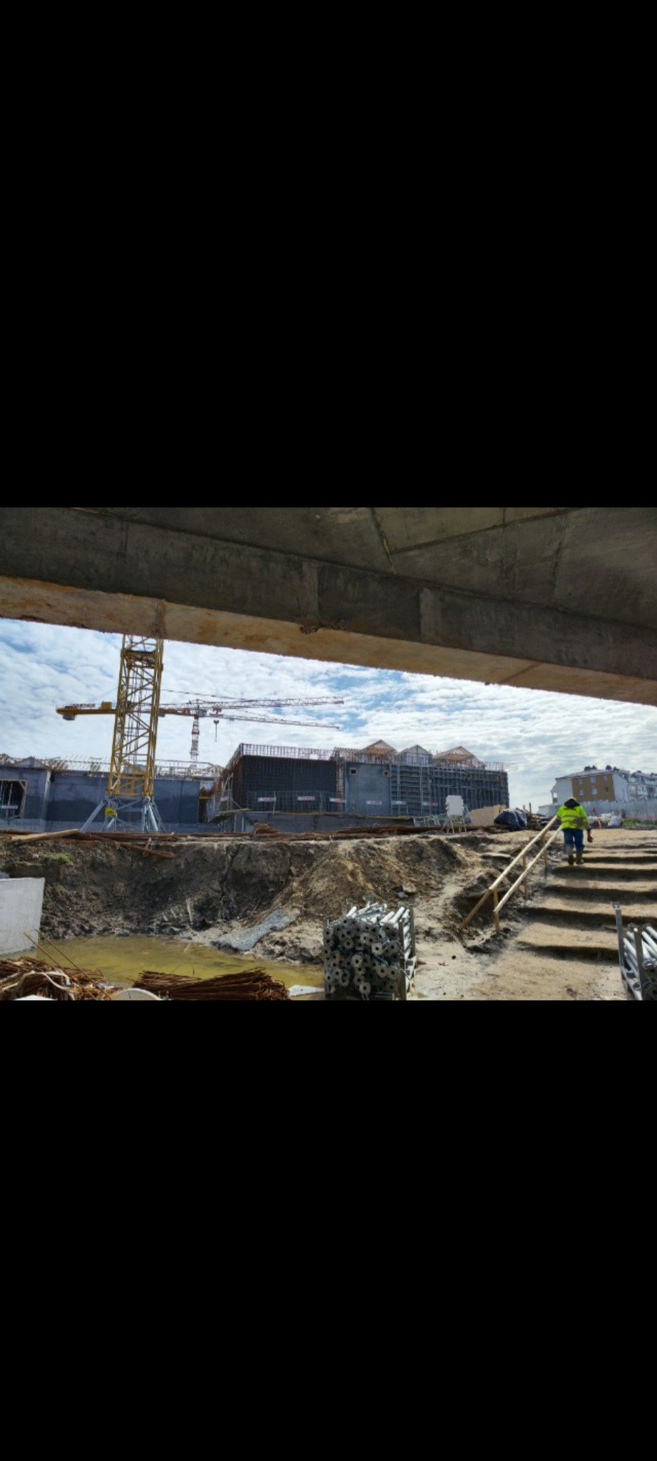 Plac budowy z widocznymi elementami konstrukcyjnymi, żurawiami i pracownikiem w odblaskowej kamizelce w tle, z ziemią i materiałami budowlanymi na pierwszym planie, ujęcie spod betonowej konstrukcji.