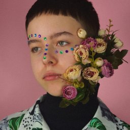 Nadia Wadelska - Portret osoby z dekoracjami z kwiatów i kolorowych kamieni na twarzy, na różowym tle.