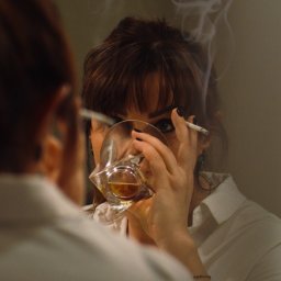 Nadia Wadelska - Kobieta w białej koszuli spogląda w lustro, trzymając w dłoni kwadratową szklankę z bursztynowym płynem i papierosa, z którego unosi się dym.
