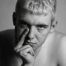 Nadia Wadelska - Czarno-biały portret młodego mężczyzny z platynowymi włosami, zamyślony wyraz twarzy, dłoń przy twarzy, na szyi cienki łańcuszek z koralików.