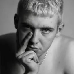 Czarno-biały portret młodego mężczyzny z platynowymi włosami, zamyślony wyraz twarzy, dłoń przy twarzy, na szyi cienki łańcuszek z koralików.