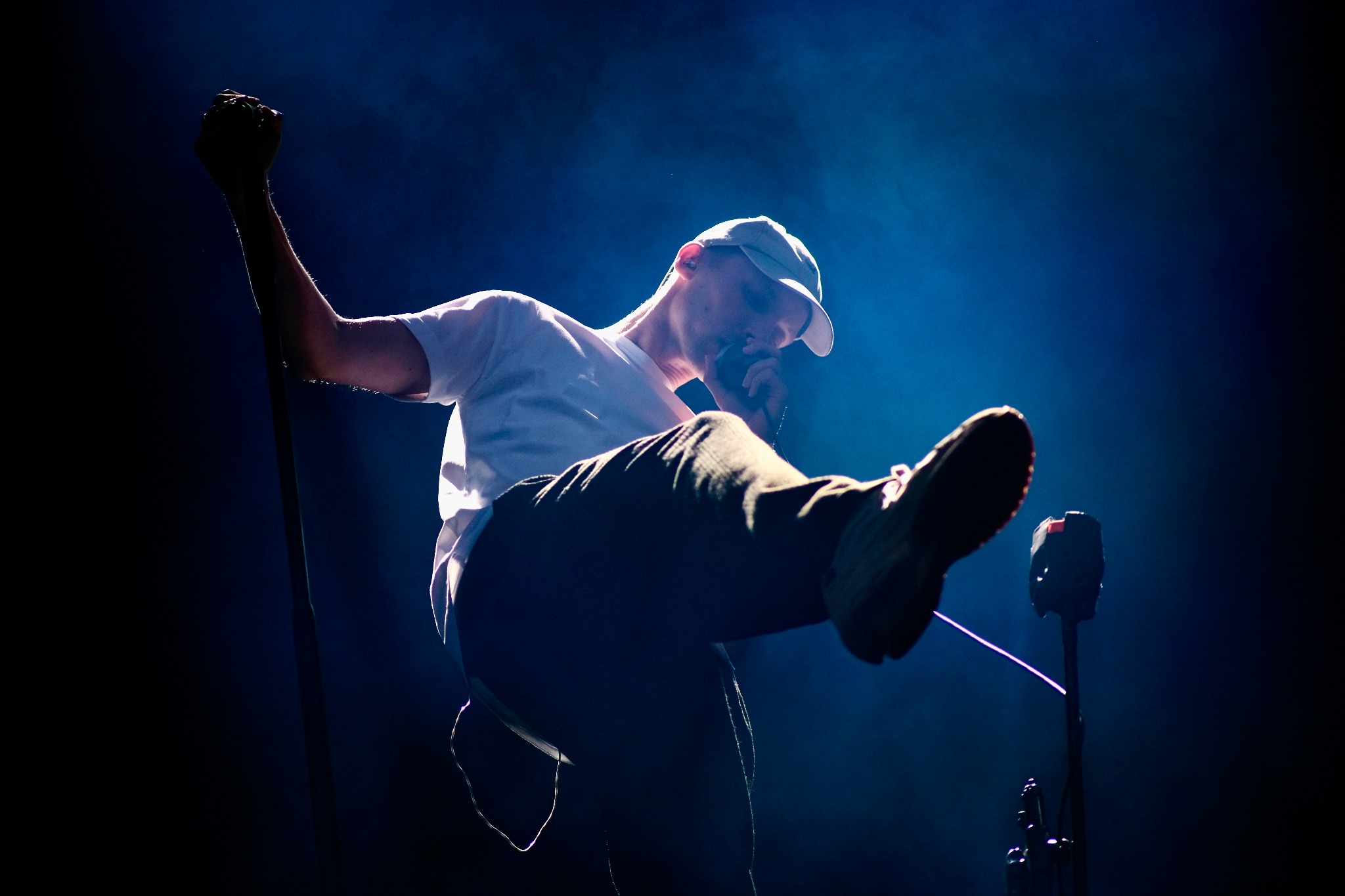 Piosenkarz z podniesioną ręką i nogą w górze, śpiewa do mikrofonu na scenie oświetlonej niebieskim światłem.