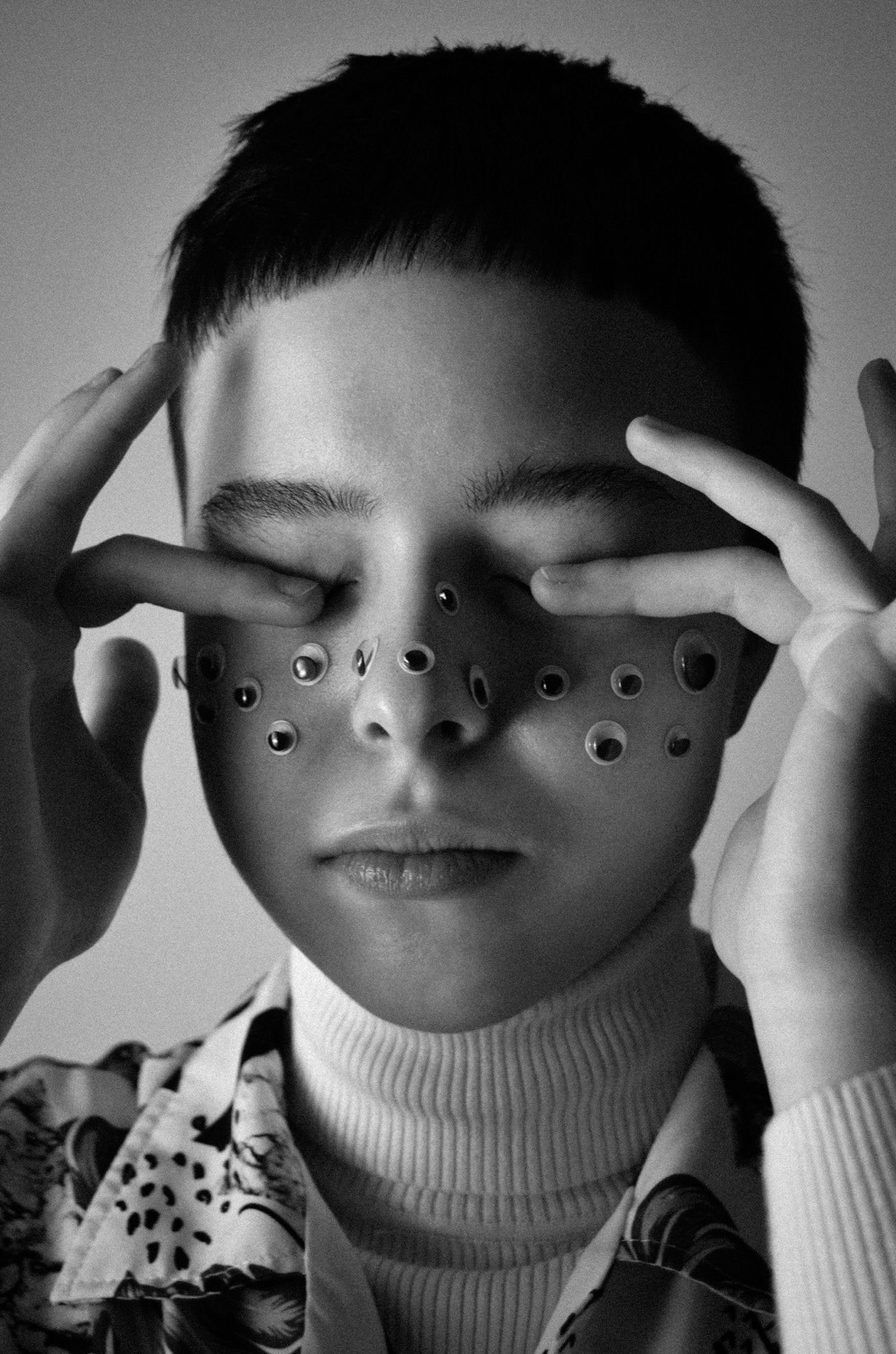 Czarno-białe zdjęcie portretowe osoby z zamkniętymi oczami i googly eyes przyklejonymi do twarzy, dotykającej skroni palcami.