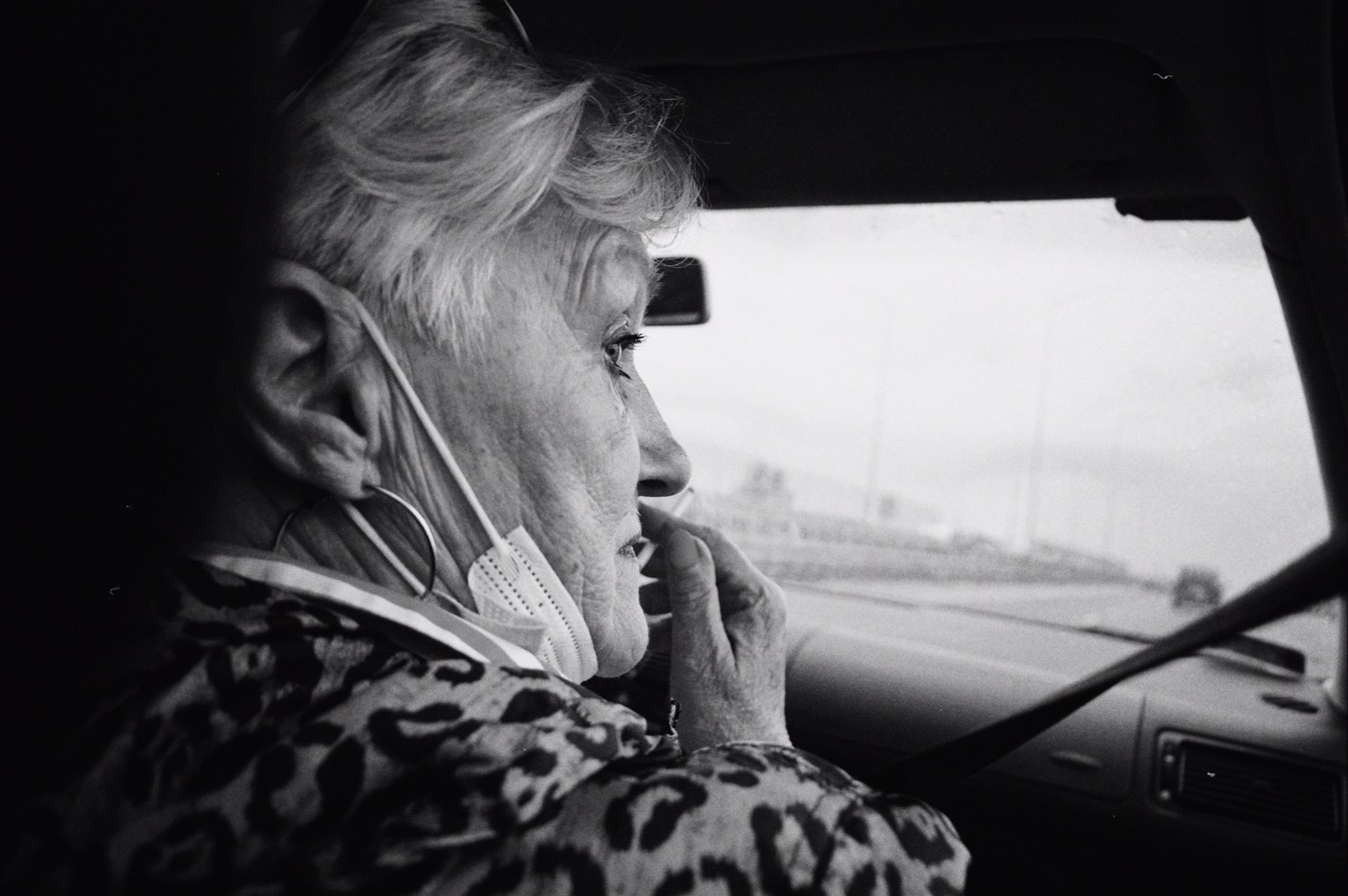 Czarno-białe zdjęcie profilu starszej kobiety w samochodzie, z maską na szyi i wzrokiem skierowanym w dal, uchwycone z perspektywy pasażera.