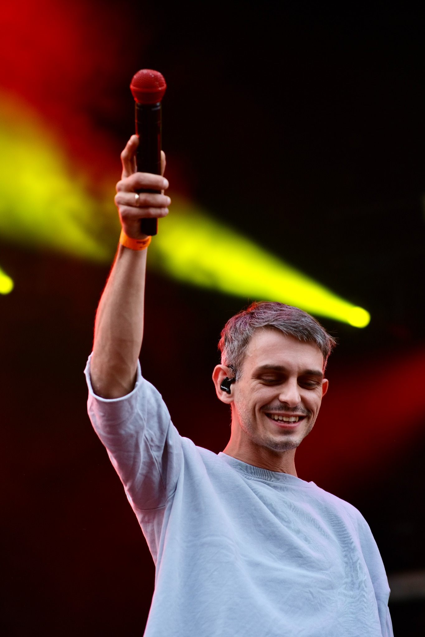 Uśmiechnięty mężczyzna w szarej bluzie trzyma uniesiony mikrofon z czerwoną główką na scenie oświetlonej czerwonym i żółtym światłem.
