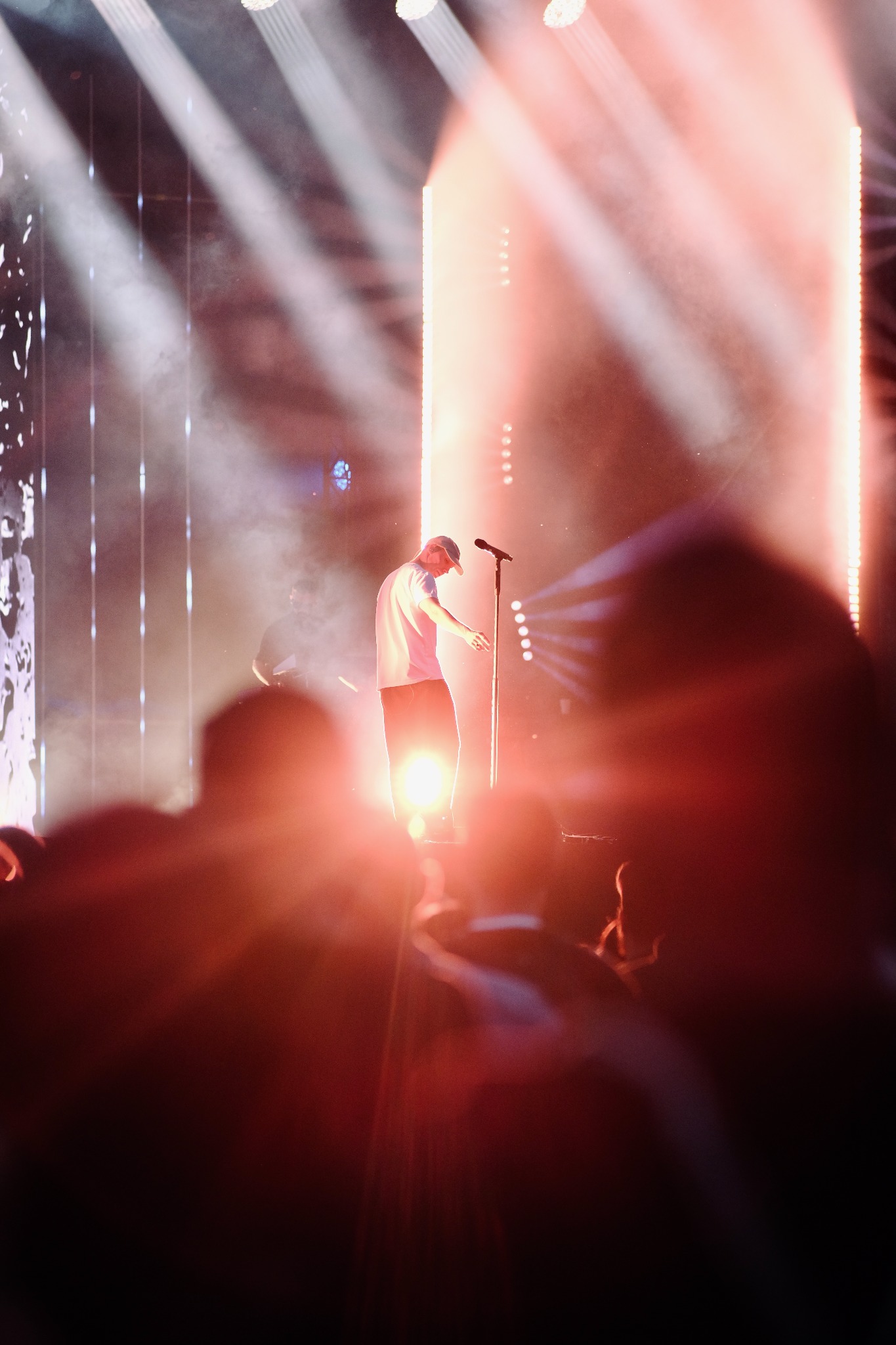 Piosenkarz w bejsbolówce śpiewa do mikrofonu na scenie oświetlonej reflektorami, widownia w tle.