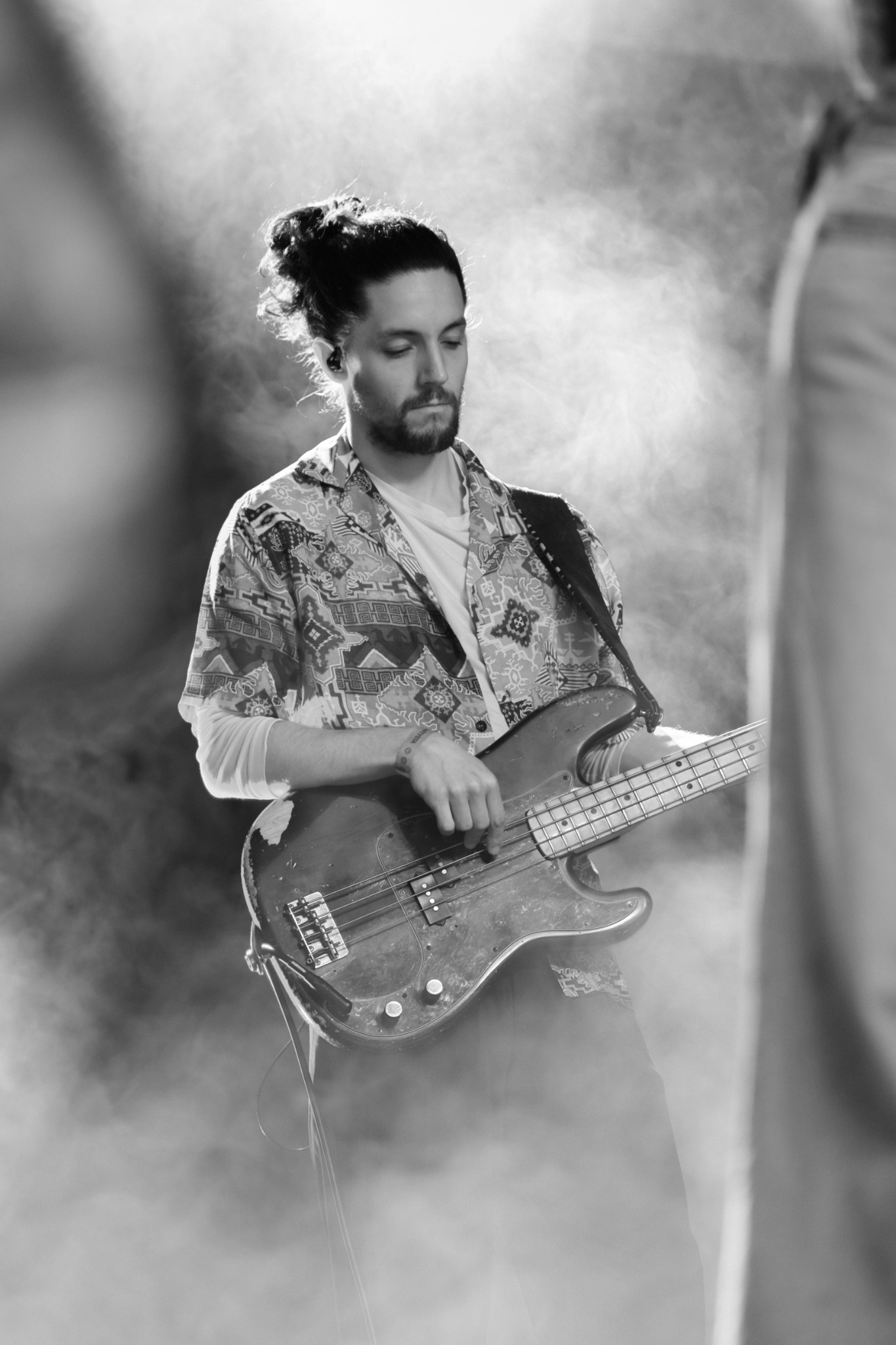 Czarno-białe zdjęcie muzyka grającego na basie na scenie w oparach dymu, z włosami spiętymi w kok, w koszuli we wzory.