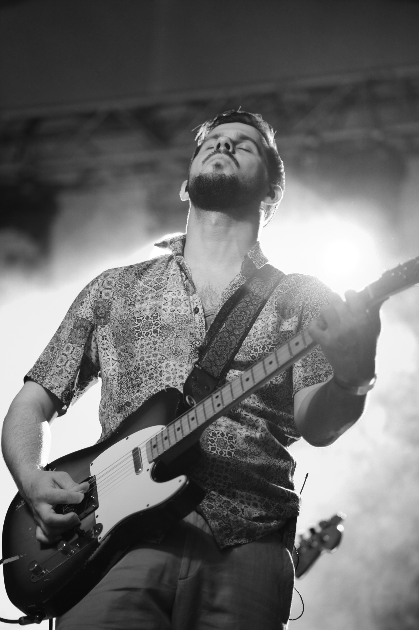 Czarno-białe zdjęcie muzyka grającego na gitarze elektrycznej z zamkniętymi oczami podczas występu na scenie z oświetleniem w tle.
