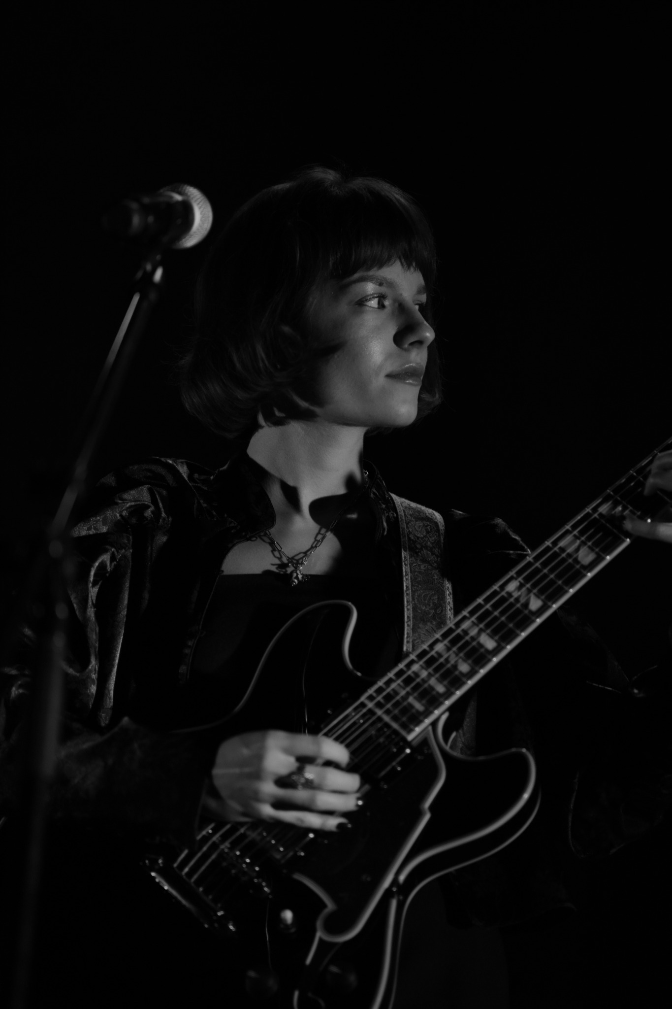 Czarno-białe zdjęcie kobiety z gitarą elektryczną na scenie, oświetlonej światłem reflektorów, z mikrofonem w tle.