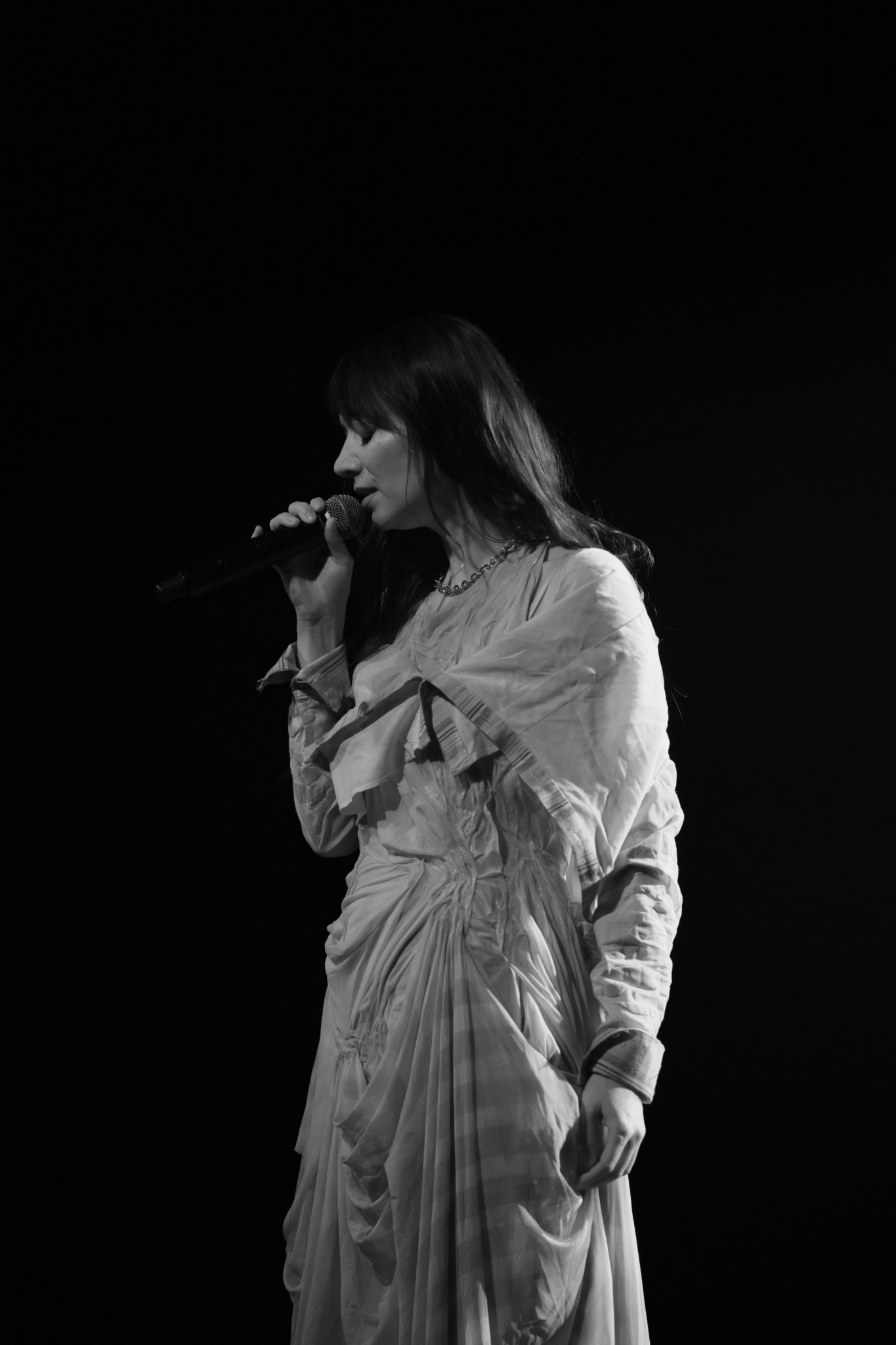 Czarno-białe zdjęcie kobiety z mikrofonem, w drapowanej sukni, oświetlonej punktowo na czarnym tle.