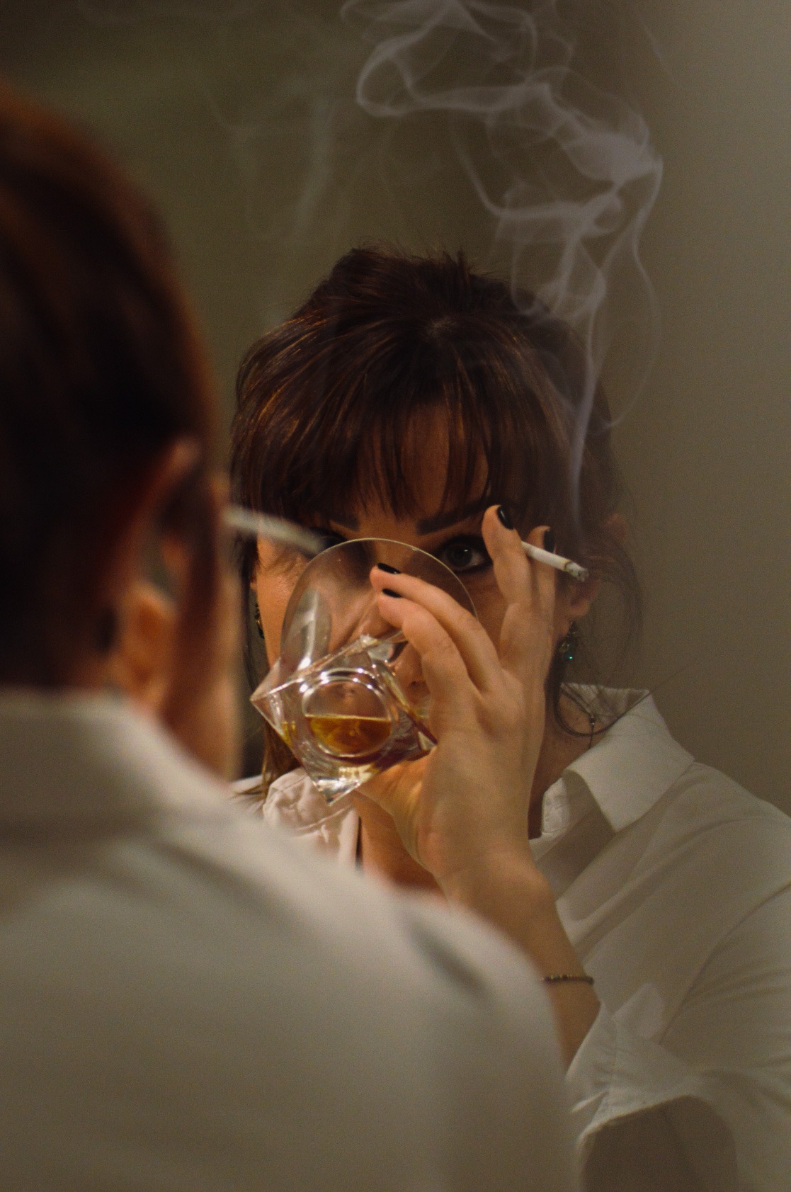 Kobieta w białej koszuli spogląda w lustro, trzymając w dłoni kwadratową szklankę z bursztynowym płynem i papierosa, z którego unosi się dym.