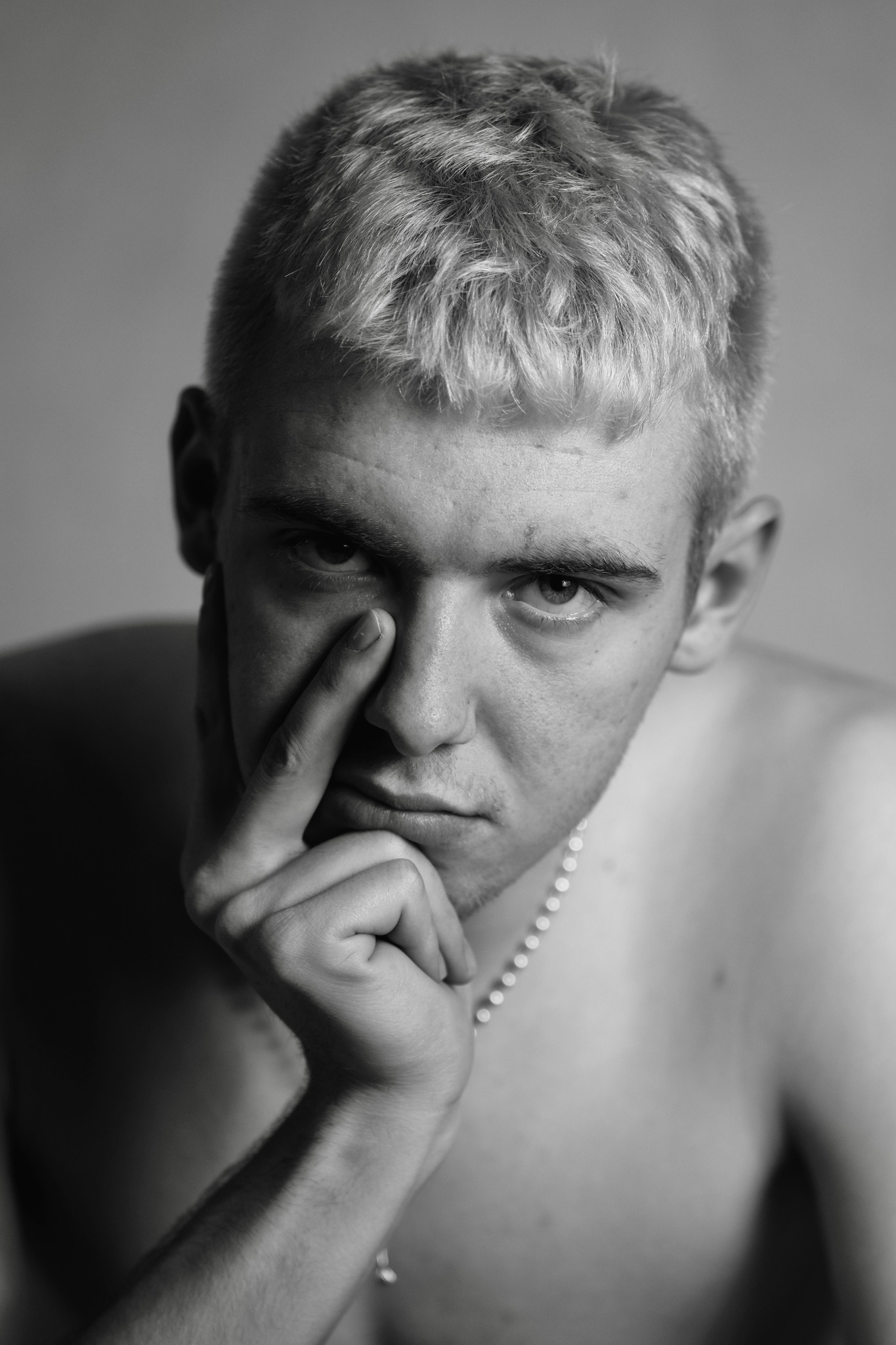 Czarno-biały portret młodego mężczyzny z platynowymi włosami, zamyślony wyraz twarzy, dłoń przy twarzy, na szyi cienki łańcuszek z koralików.