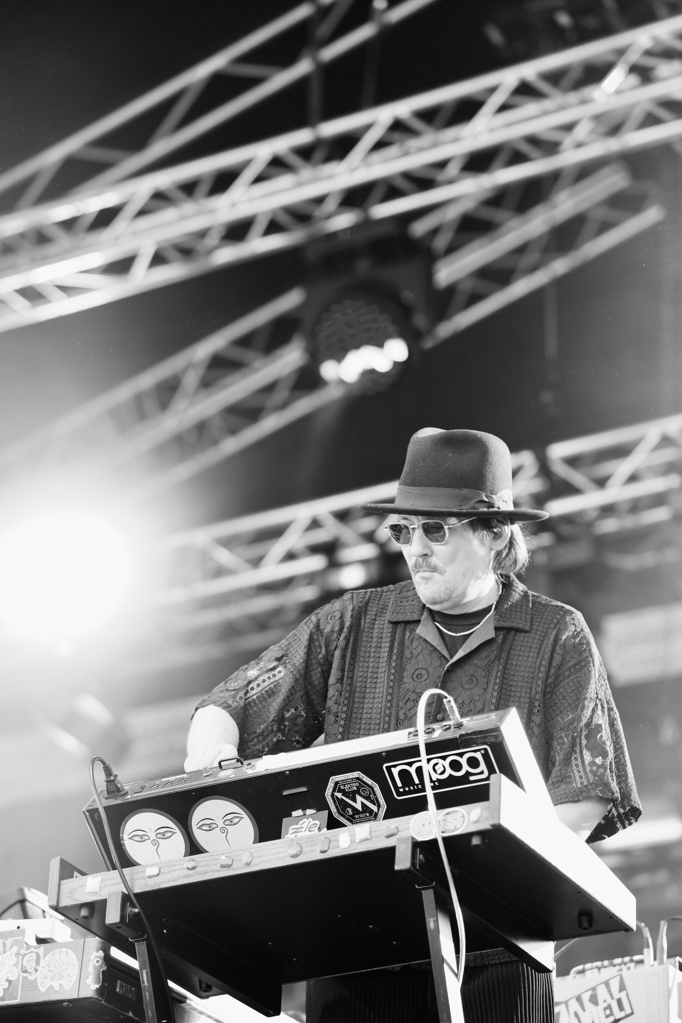 Czarno-białe zdjęcie muzyka w kapeluszu i okularach, grającego na syntezatorze Moog na scenie z metalową konstrukcją w tle.