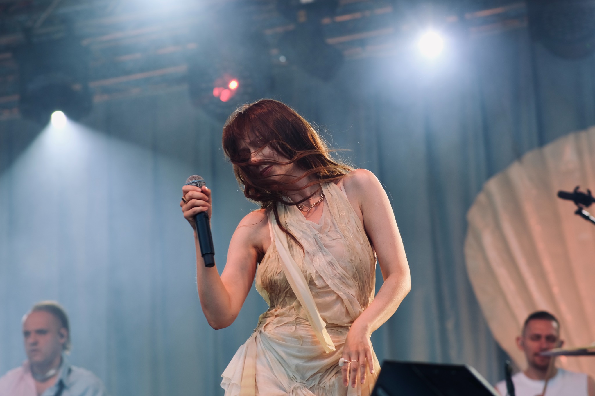 Wokalistka z rozwianymi włosami śpiewa na scenie w zwiewnej sukience, trzymając mikrofon, w tle widoczni muzycy i reflektory.