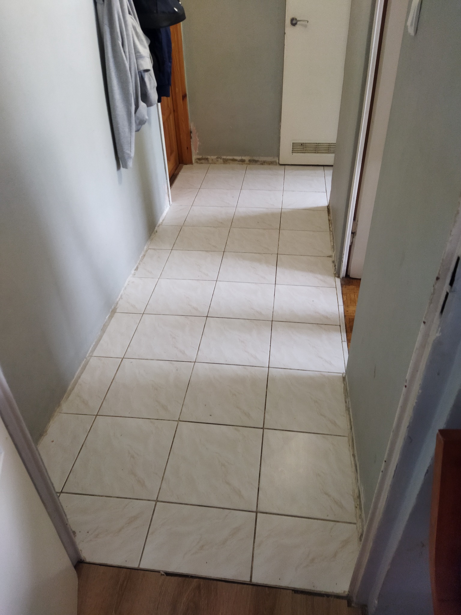 Korytarz wyłożony kwadratowymi, beżowymi płytkami podłogowymi z widocznymi fugami, z szarymi ścianami i dwiema parami drzwi po bokach, gdzie widać fragment drewnianej podłogi w przejściu.