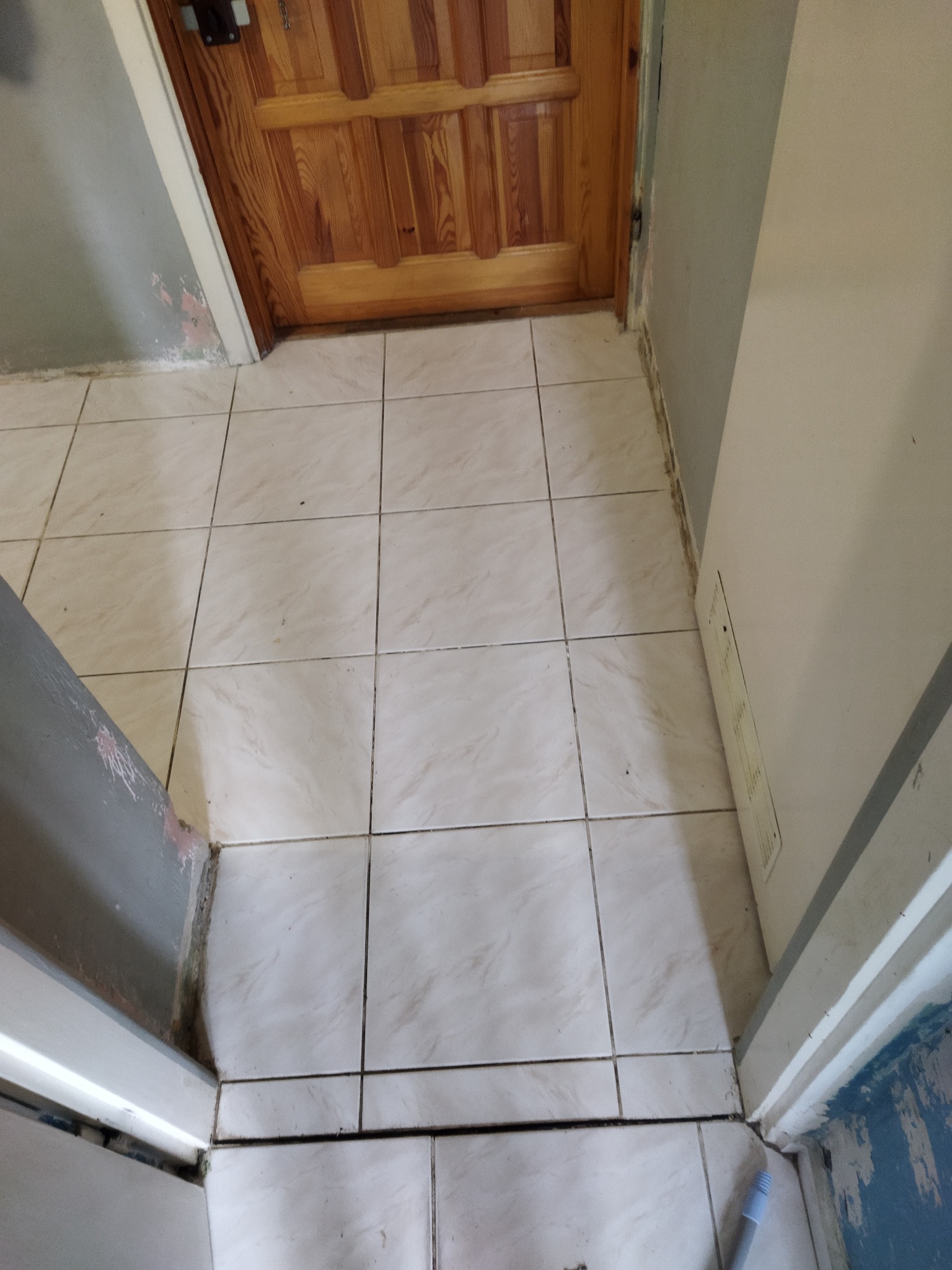 Beżowe płytki podłogowe ułożone w kwadraty, widoczne przy drewnianych drzwiach i ścianie z śladami farby, ukazujące stan przed remontem.