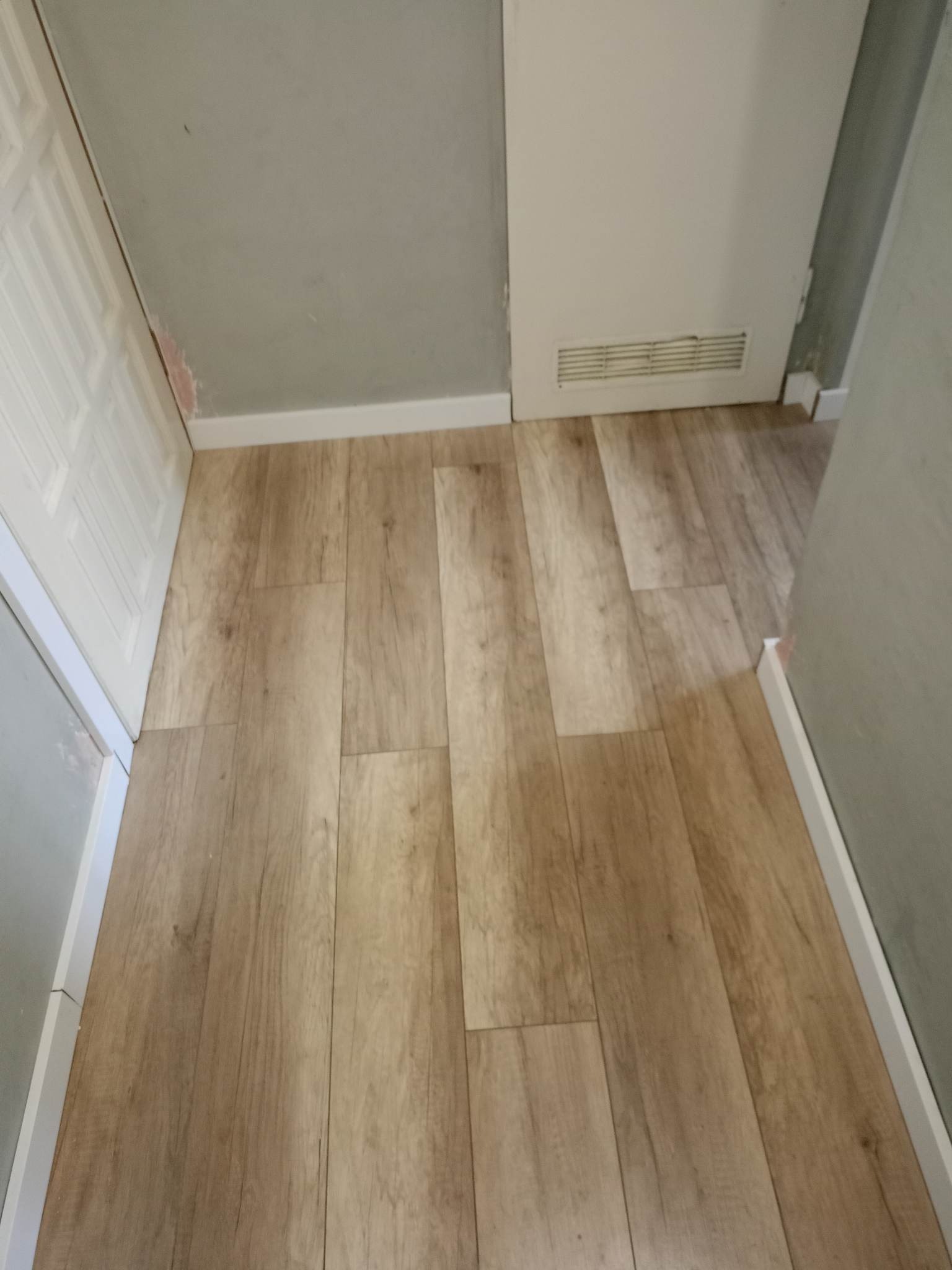 Ułożona podłoga z paneli imitujących drewno, widoczne białe listwy przypodłogowe, fragment ściany w szarym odcieniu i białe drzwi z widocznymi śladami po gładzi.