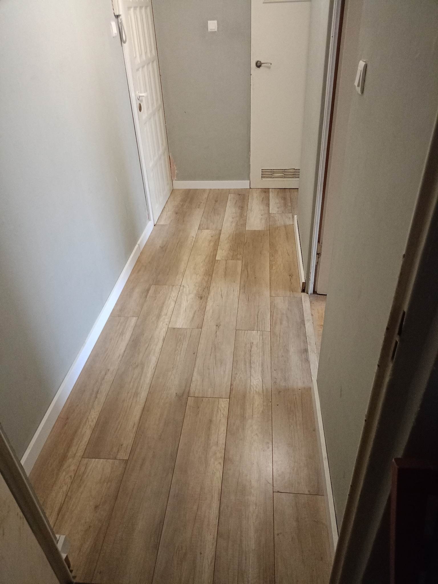 Korytarz z ułożoną podłogą imitującą drewno, białe listwy przypodłogowe, szare ściany i dwie białe drzwi.