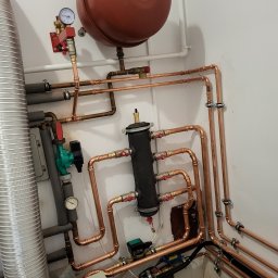 City Project Sp. z o.o. - Kompleksowa instalacja hydrauliczna z miedzianych rur, widoczny zbiornik wyrównawczy, pompa obiegowa, wymiennik ciepła i manometry, całość zamontowana przy białej ścianie.