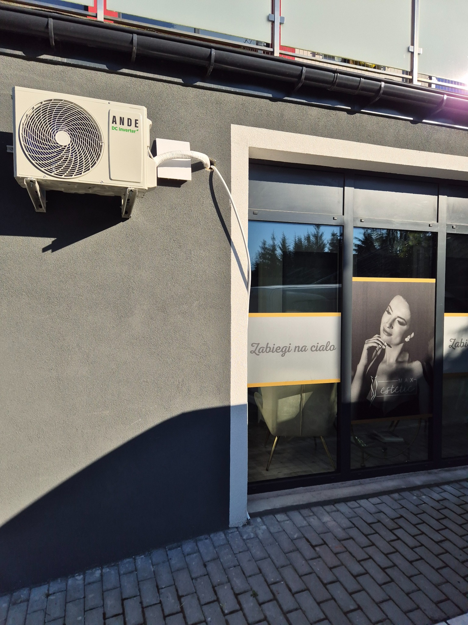 Zamontowana jednostka zewnętrzna klimatyzacji ANDE DC Inverter na szarej elewacji budynku z oknami ozdobionymi napisem 'Zabiegi na ciało' i portretem kobiety.