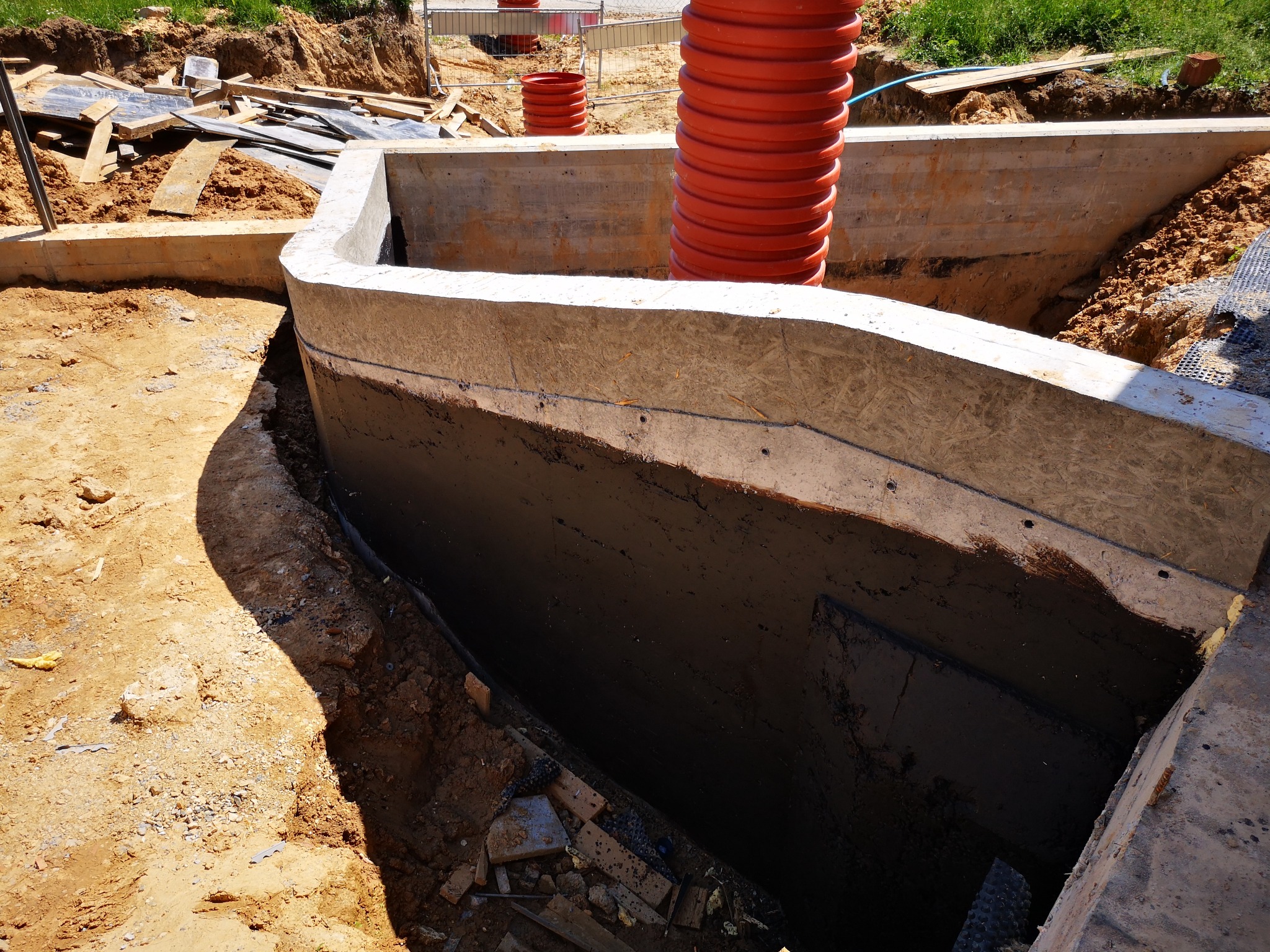 Zabezpieczona fundamentowa ściana betonowa w wykopie, pomalowana czarną izolacją, z pomarańczową rurą wentylacyjną i deskami w tle.