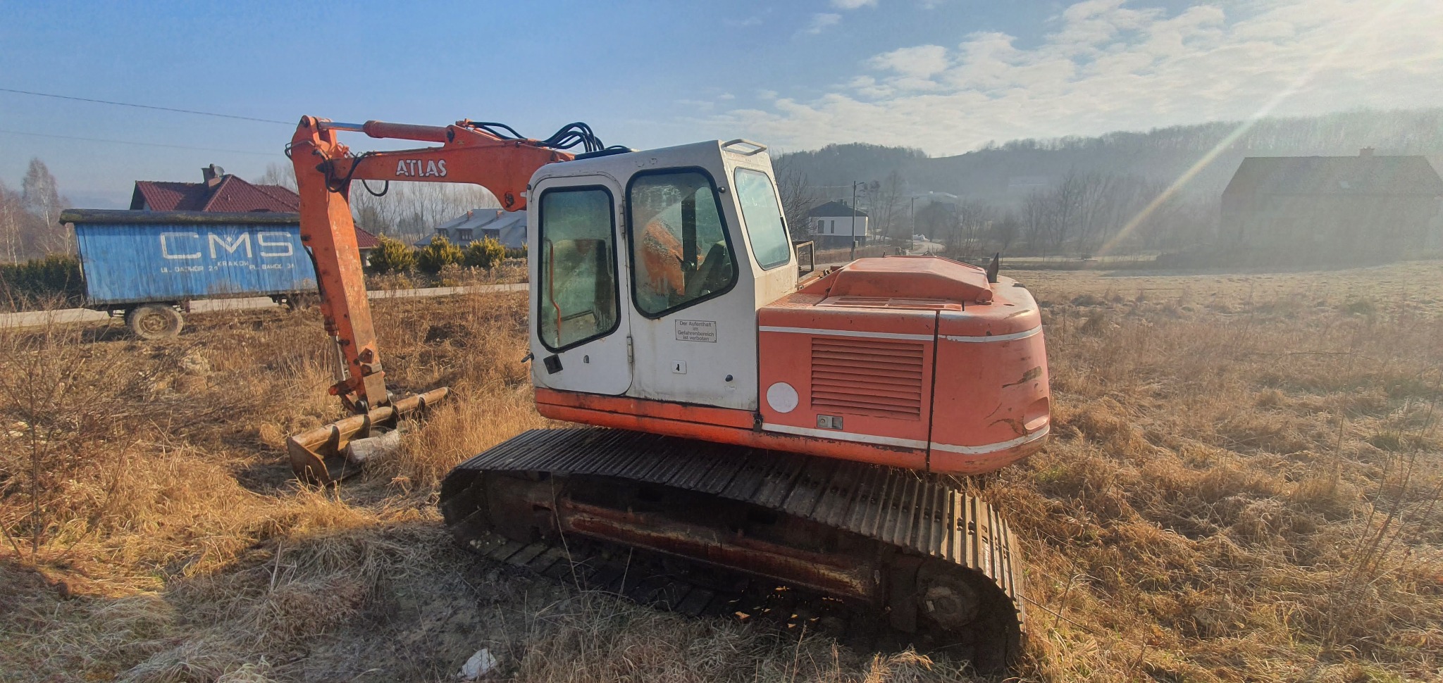 Pomarańczowy, używany koparka gąsienicowa Atlas na zarośniętym polu, z przyczepą CMS w tle i zalesionym wzgórzem w oddali.
