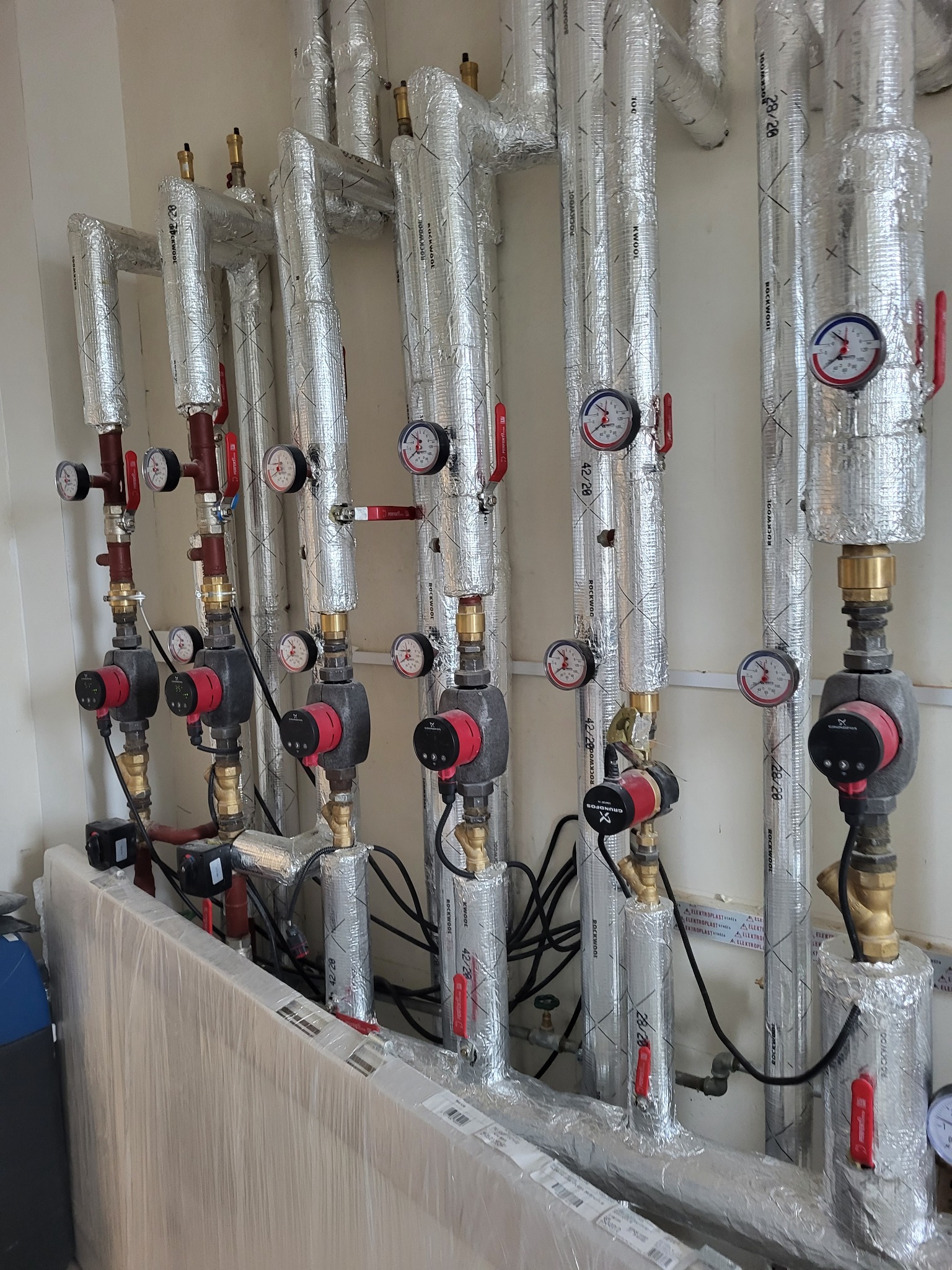 Rzędy pionowych, zaizolowanych rur instalacji grzewczej z manometrami i pompami obiegowymi Grundfos, zamontowane przy białej ścianie. Część rur owinięta folią aluminiową z widocznymi napisami...
