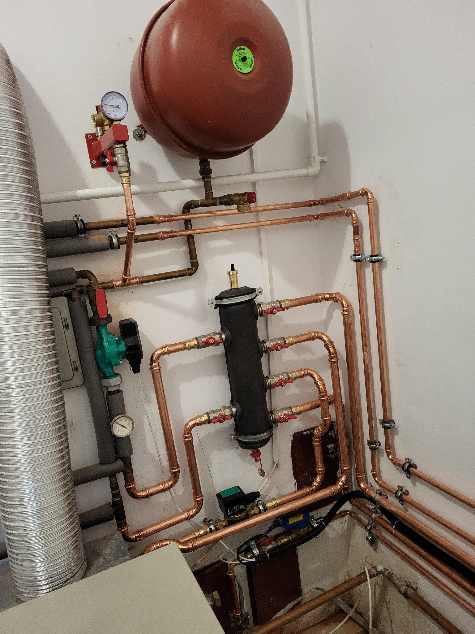 Kompleksowa instalacja hydrauliczna z miedzianych rur, widoczny zbiornik wyrównawczy, pompa obiegowa, wymiennik ciepła i manometry, całość zamontowana przy białej ścianie.