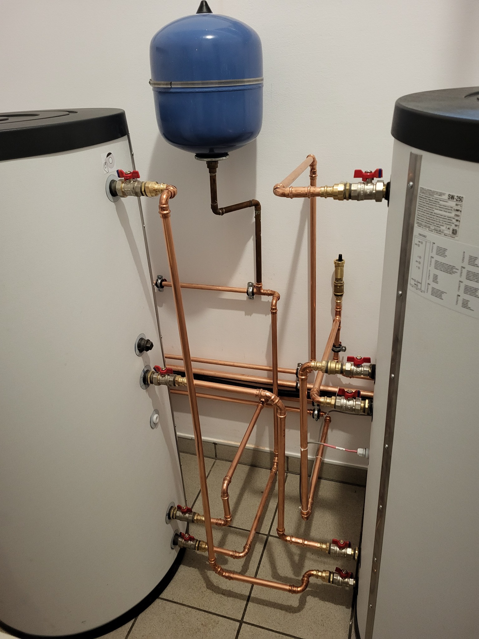 Instalacja hydrauliczna z miedzianymi rurami i zaworami, podłączona do dwóch zbiorników ciepłej wody i niebieskiego naczynia wzbiorczego, widoczna na tle białej ściany i płytek podłogowych.