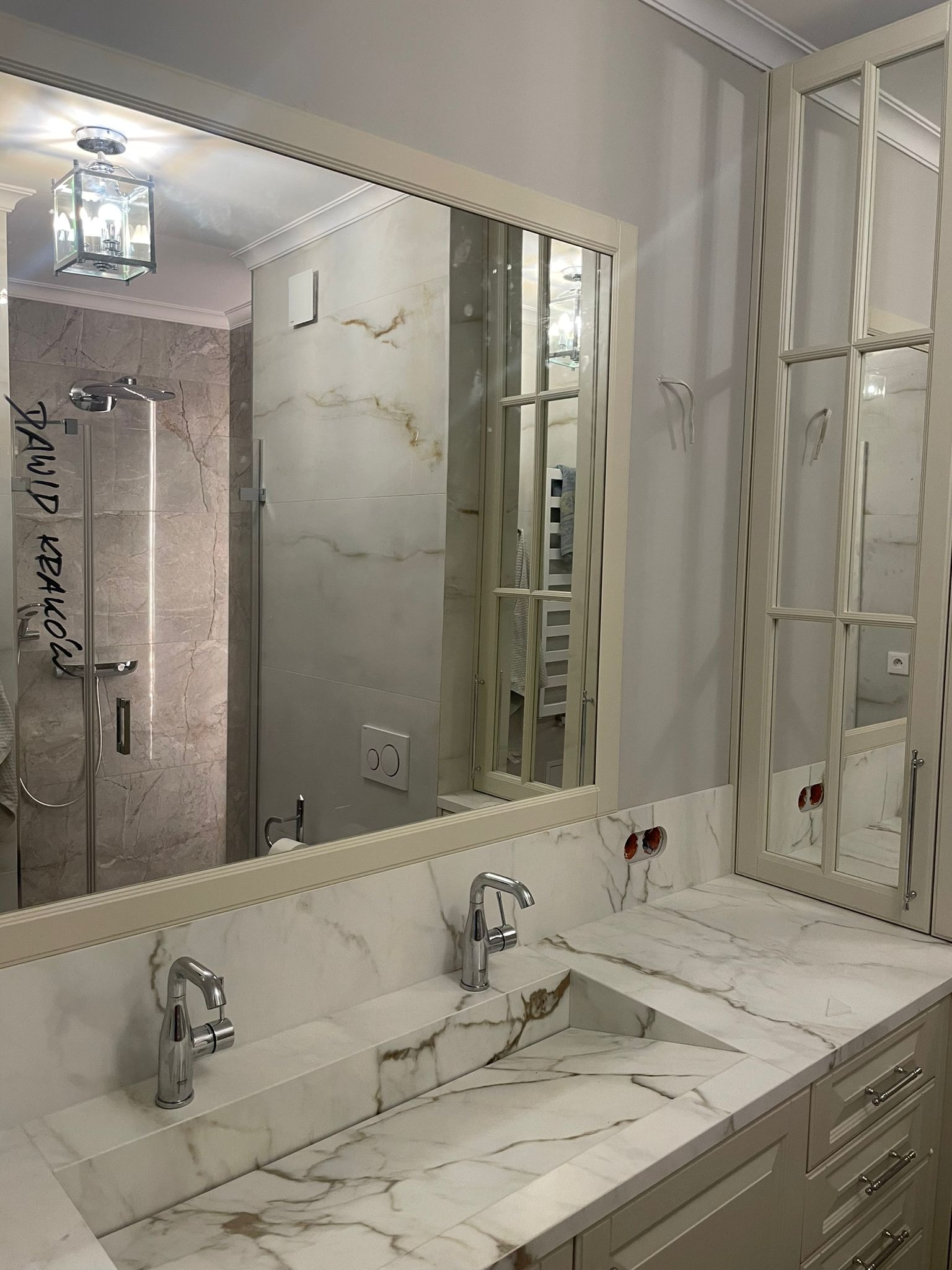 Elegancka łazienka z marmurowym blatem umywalki, lustrem z widocznym odbiciem kabiny prysznicowej i szafkami w kolorze złamanej bieli, widoczne gniazdka elektryczne bez osprzętu.