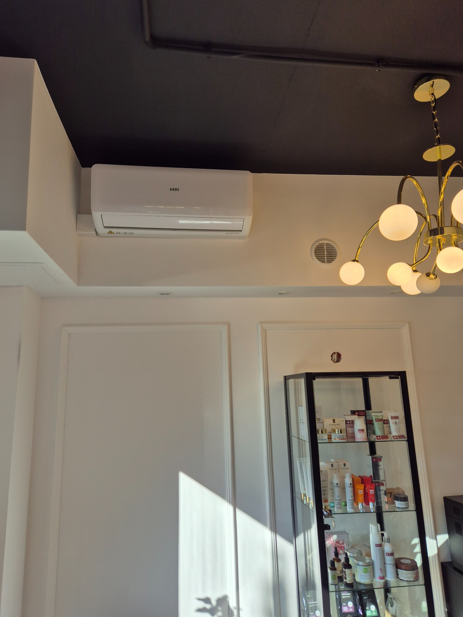 Biała klimatyzacja marki Ande zamontowana na ścianie w pomieszczeniu z czarnym sufitem, widoczny ozdobny żyrandol i przeszklona witryna z kosmetykami.