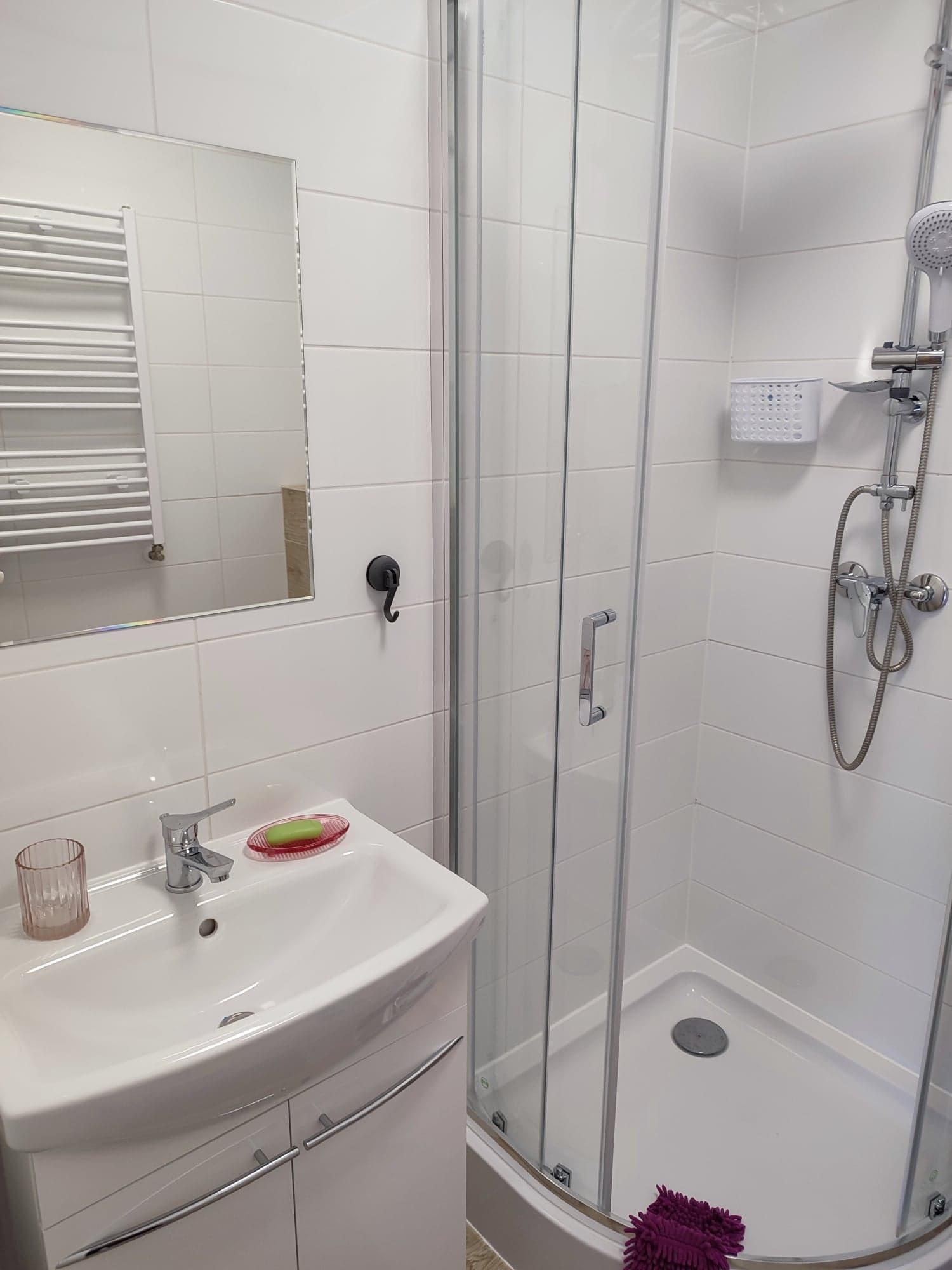 Nowoczesna łazienka z białą umywalką z szafką, kabiną prysznicową z przezroczystymi drzwiami i grzejnikiem drabinkowym widocznym w lustrze.