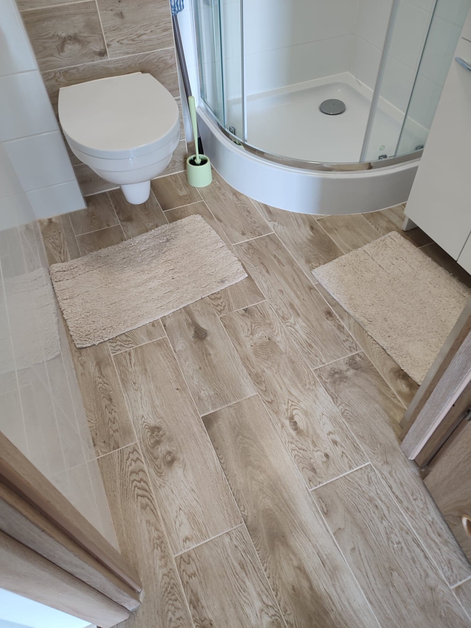 Nowoczesna łazienka z kabiną prysznicową, toaletą podwieszaną, drewnopodobnymi płytkami podłogowymi i beżowymi dywanikami.