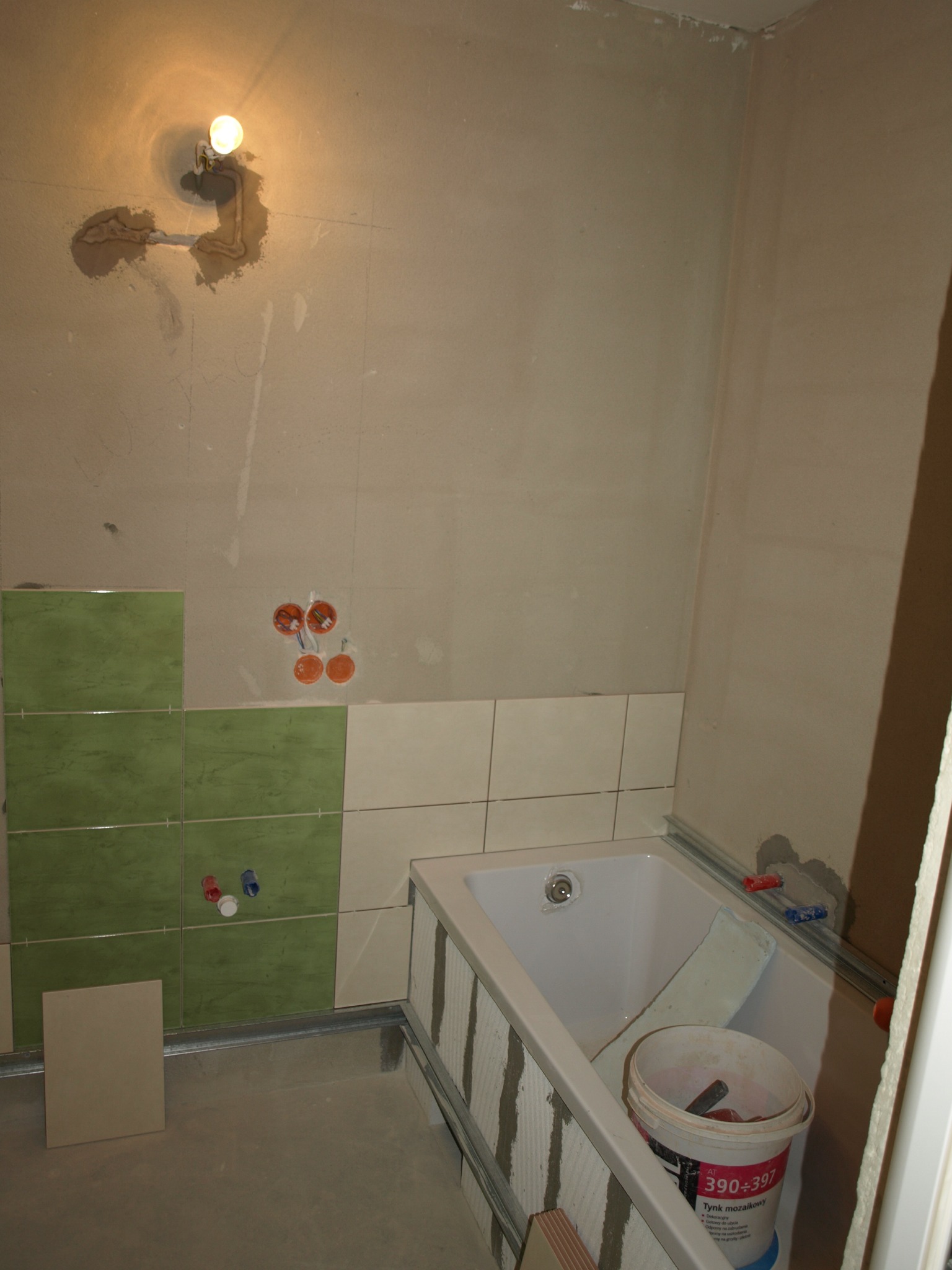 Łazienka w trakcie remontu: częściowo położone zielone i beżowe płytki, widoczne instalacje wodne i elektryczne, wanna z elementami mocującymi, wiadro z zaprawą mozaikową.