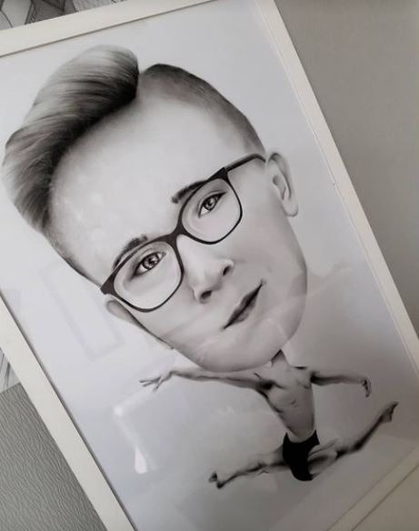 Portret karykaturalny w czerni i bieli, przedstawiający mężczyznę w okularach z ciałem tancerza baletowego, oprawiony w białą ramkę.