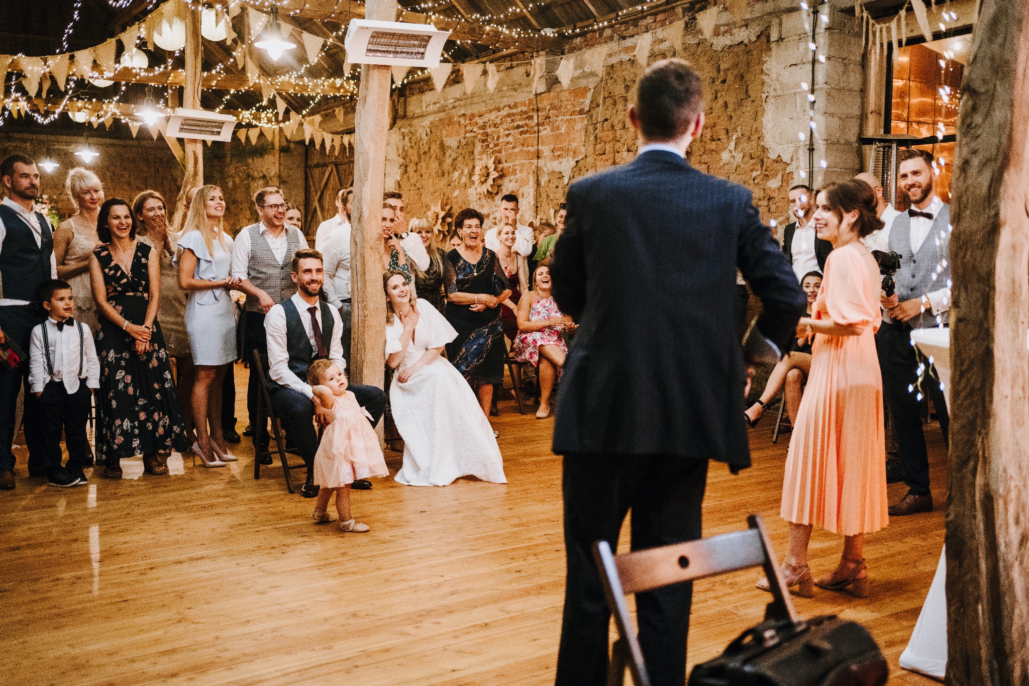 Iluzjonista prezentuje pokaz na weselu. Goście weselni obserwują z zaciekawieniem. W tle ceglana ściana i dekoracyjne lampki. Na pierwszym planie para w sukni ślubnej.