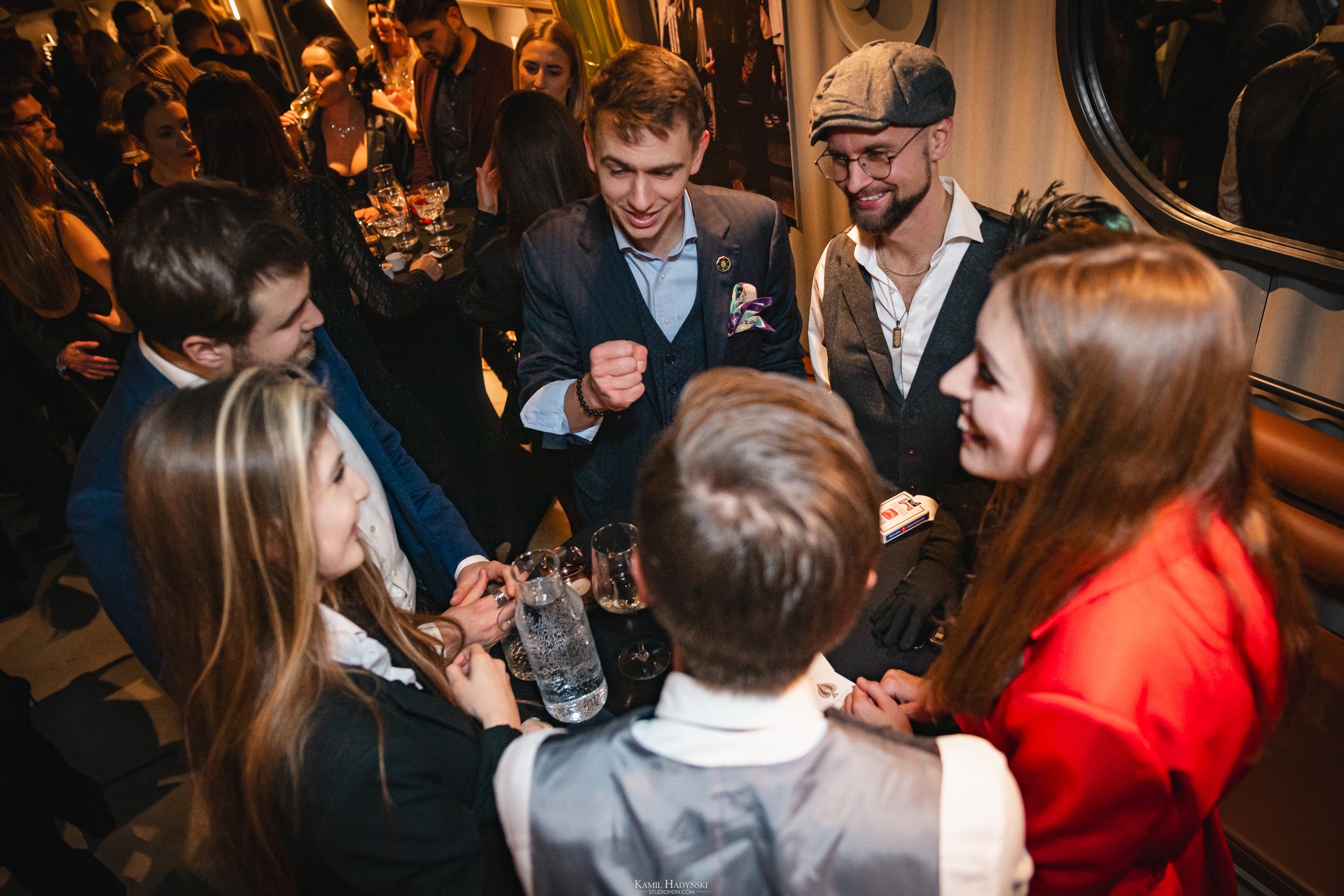 Uśmiechnięty iluzjonista w szarej marynarce prezentuje trik karciany grupie gości w eleganckim wnętrzu. Widoczna butelka wody, szklanki i talia kart.