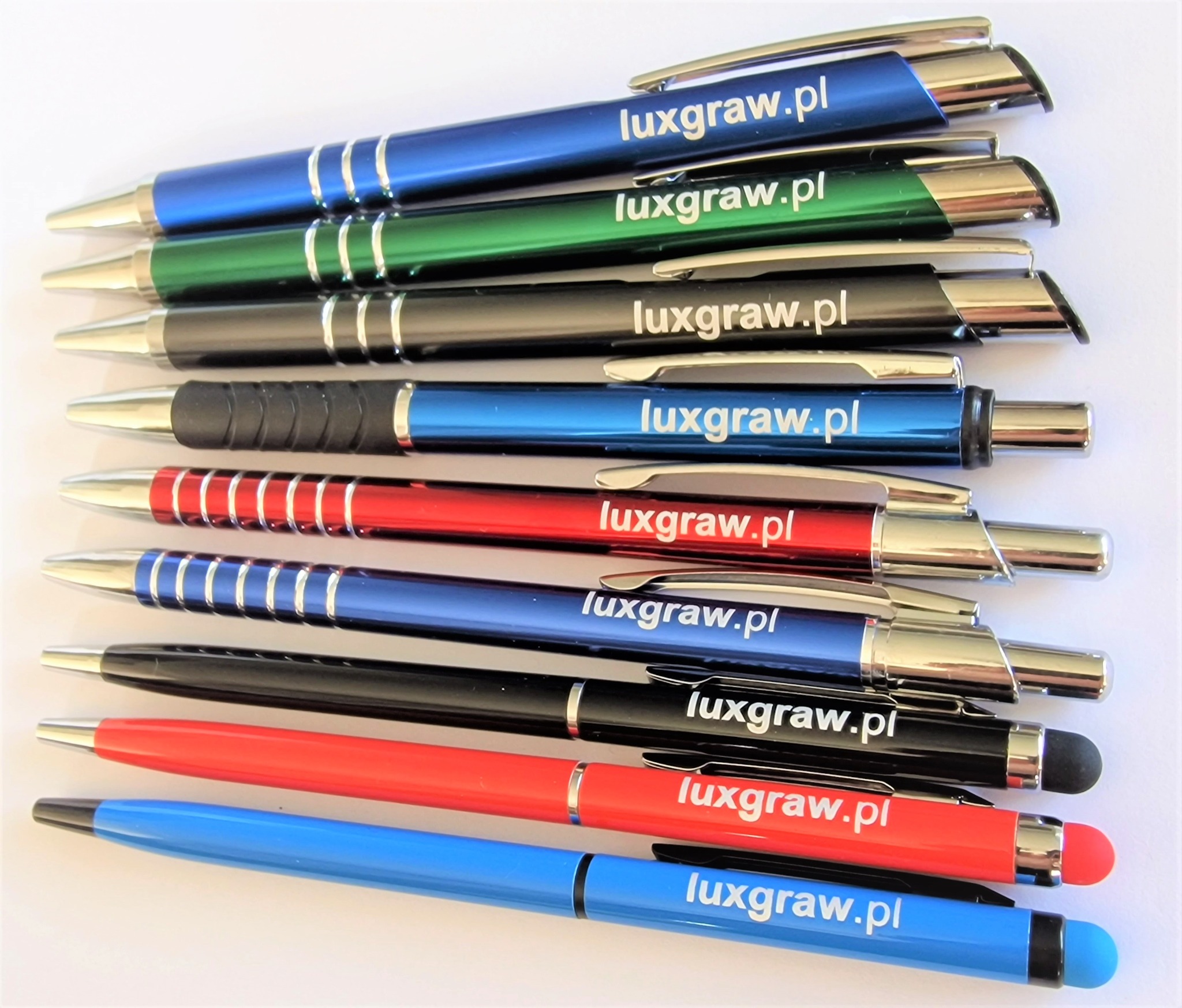 Kolekcja długopisów w różnych kolorach (niebieski, zielony, czarny, czerwony) z nadrukowanym adresem strony internetowej luxgraw.pl, niektóre z końcówką do ekranów dotykowych.