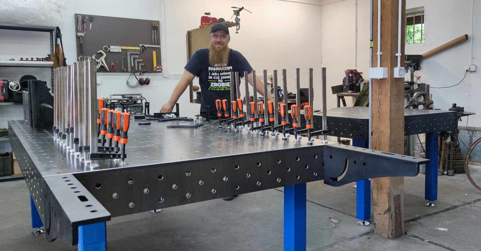 Mężczyzna z brodą w warsztacie stoi za metalowym stołem spawalniczym z otworami, na którym zamocowane są pionowe rury za pomocą pomarańczowych ścisków stolarskich. W tle narzędzia na ścianie i inne...