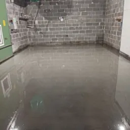 Świeżo wylana, mokra, ciemnoszara posadzka w pomieszczeniu z szarymi, ceglanymi ścianami. Na ścianie widoczny zegar i instalacje.