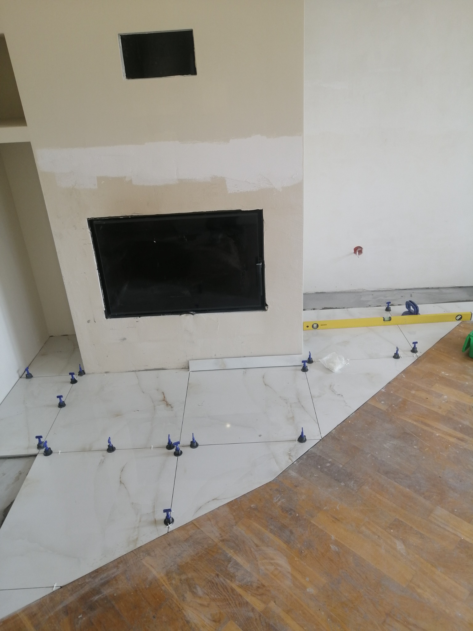 Układanie białych płytek podłogowych z imitacją marmuru, poziomica na podłodze, widoczne krzyżyki dystansowe i fragment parkietu.