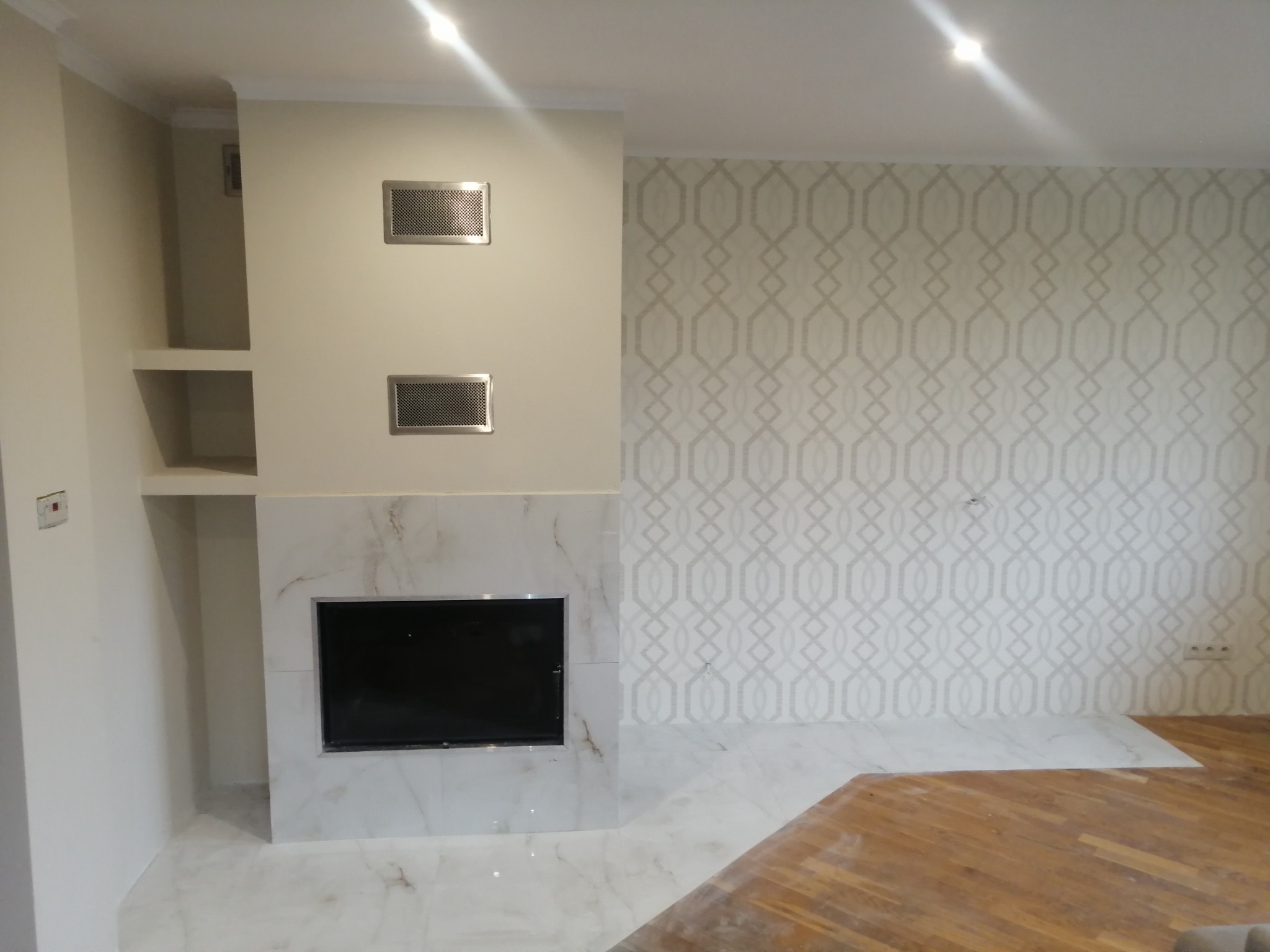 Wykończony salon z marmurowym kominkiem, ażurowymi kratkami wentylacyjnymi i tapetą w geometryczne wzory; podłoga częściowo wyłożona płytkami, częściowo drewniana w trakcie układania.