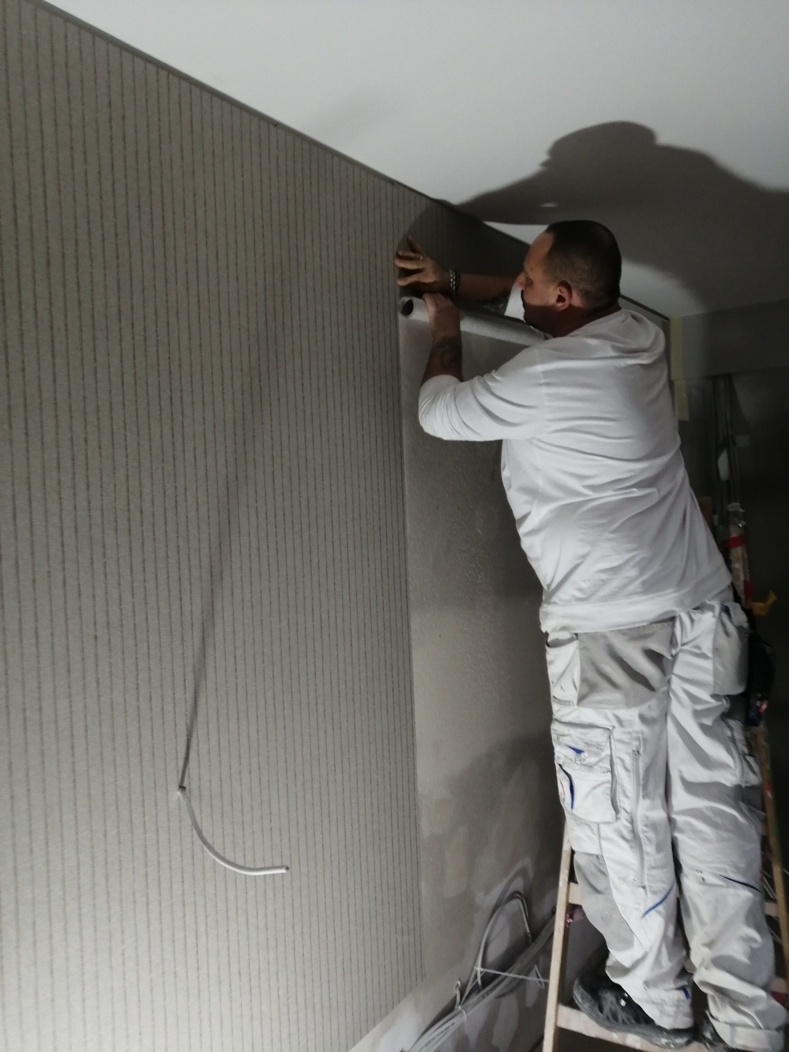 Monter na drabinie przykleja tapetę w pionowe paski do ściany, widoczny fragment wystającego kabla.