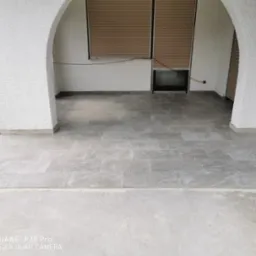 Kładzenie szarych płytek na tarasie z białymi, łukowatymi kolumnami, widoczne plastikowe krzyżyki dystansowe między płytkami, w tle brązowe rolety.