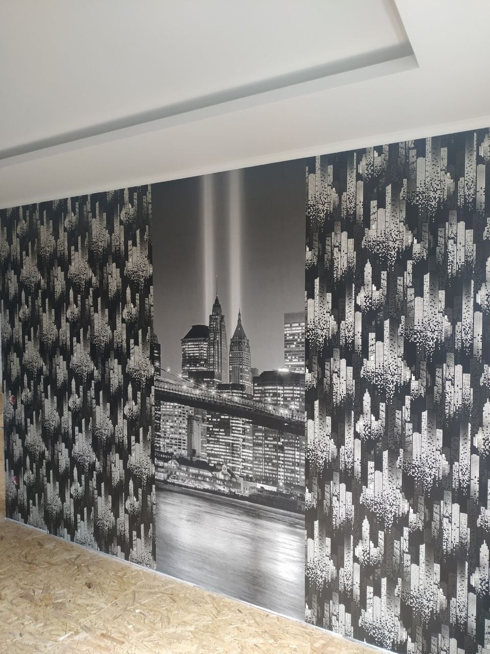 Tapeta z motywem panoramy miasta w czerni i bieli, po bokach wzór abstrakcyjnych budynków, surowa podłoga z płyt OSB.