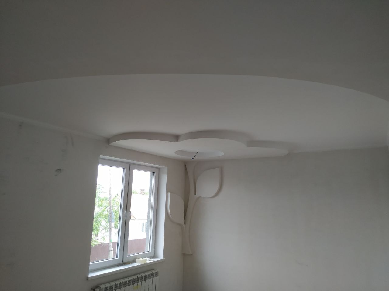 Niedokończone białe wnętrze z dekoracyjnym sztukateryjnym elementem w kształcie drzewa na ścianie obok okna i podwieszonym sufitem z owalnym wycięciem.