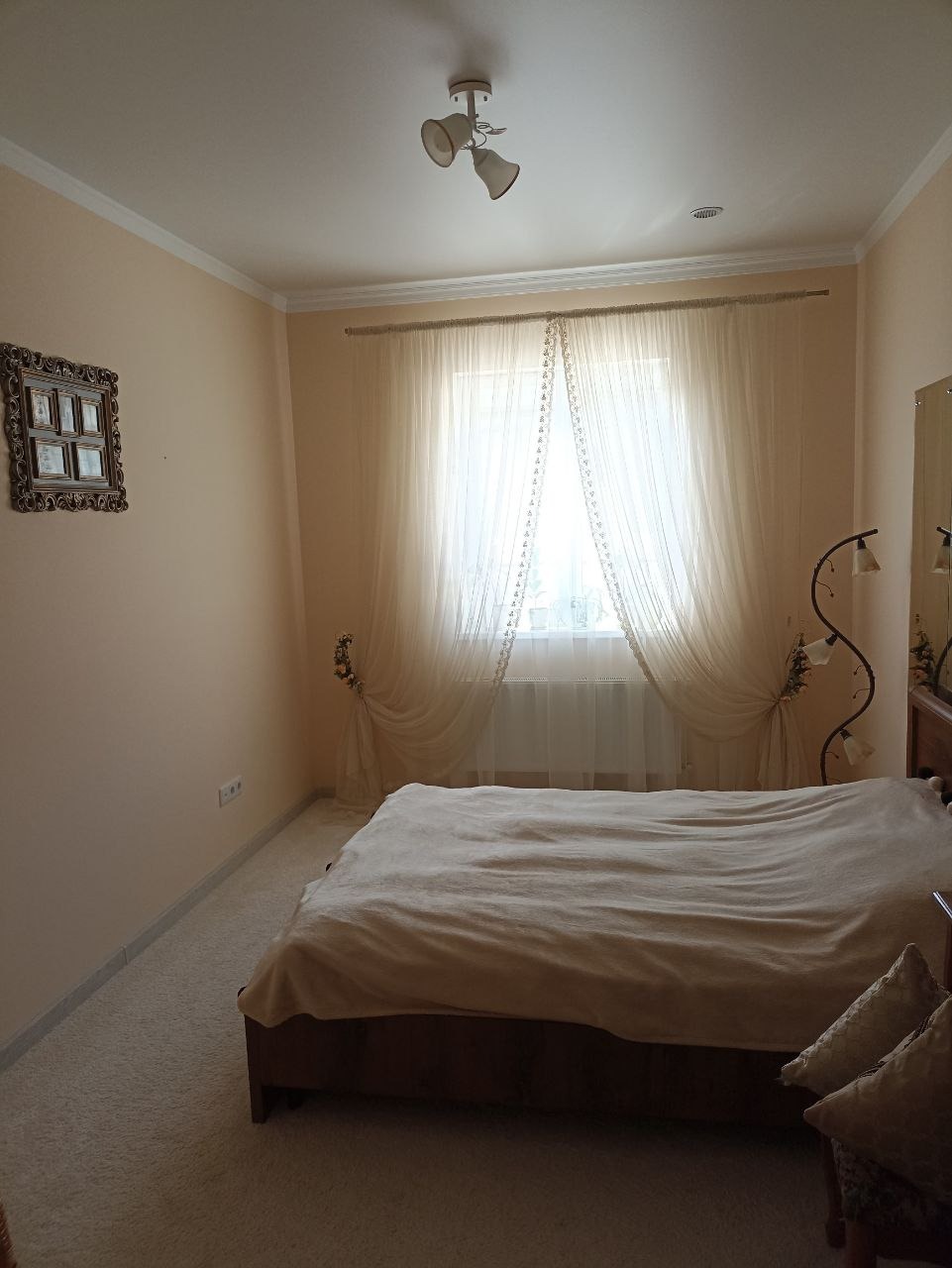 Jasna sypialnia z beżową wykładziną, łóżkiem z narzutą i dekoracyjnymi zasłonami, oświetlona lampą sufitową i lampką nocną z kwiatowym motywem.