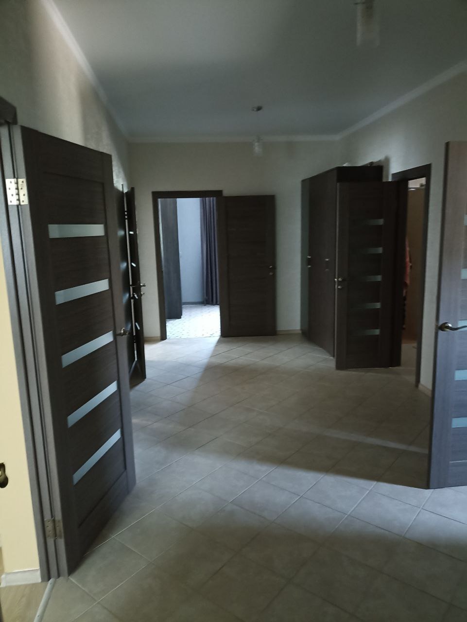 Korytarz z szarymi płytkami podłogowymi i ciemnymi drzwiami z jasnymi, poziomymi paskami, widok z perspektywy wejścia.