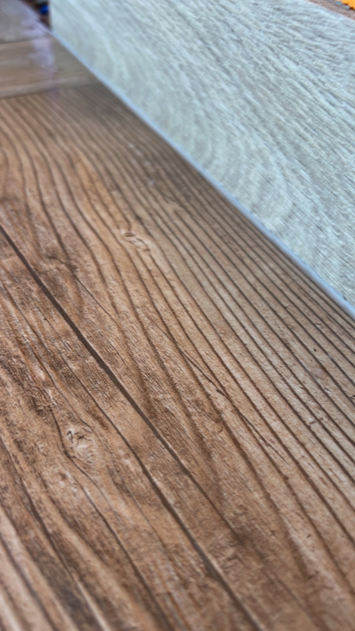 Ułożona brązowa podłoga z paneli imitujących drewno, widoczny fragment szarego panelu na ścianie w tle.