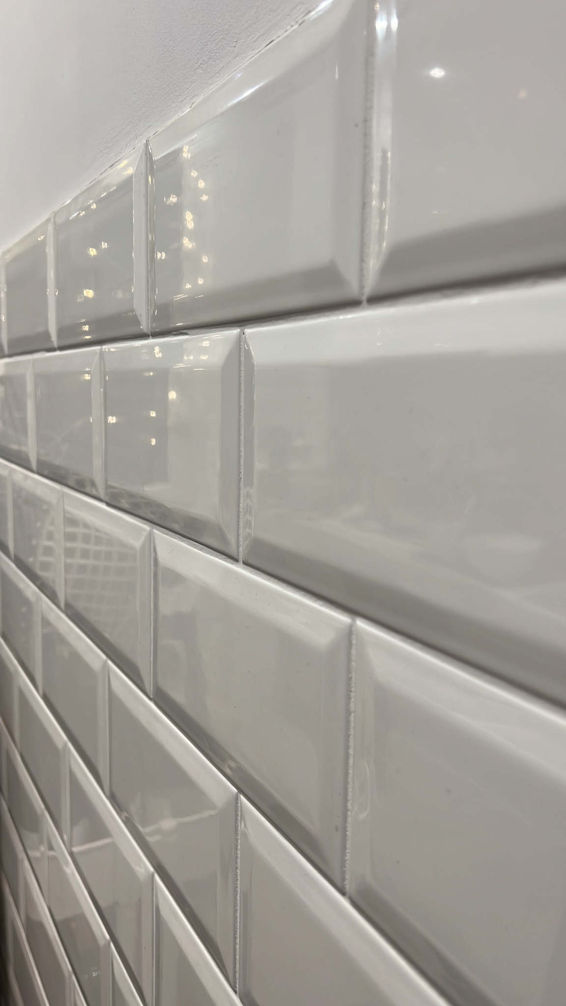 Ściana wyłożona błyszczącymi, białymi płytkami typu metro z fazowanymi krawędziami, ułożonymi w cegiełkę.